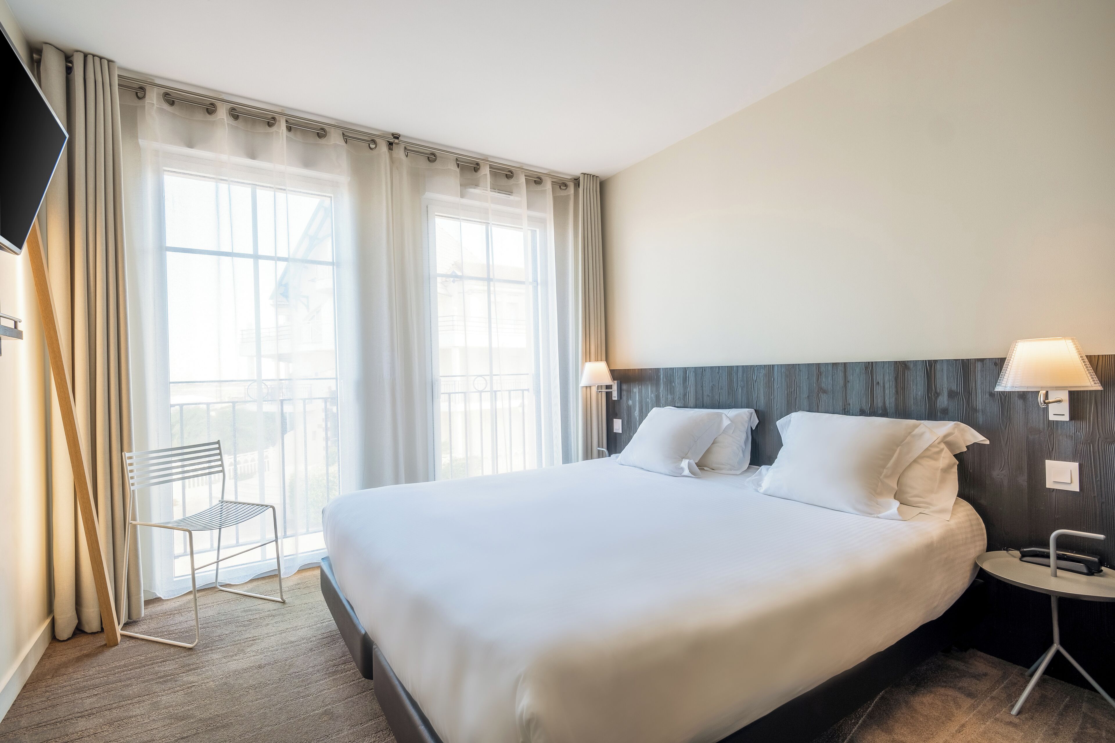 Une chambre d’hôtel avec un grand lit, deux lampes de chevet, une chaise et une fenêtre avec des rideaux.
