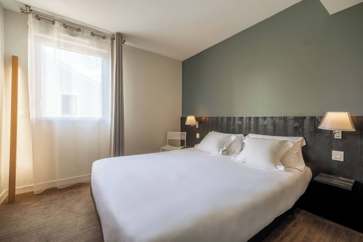 Una cama bien tendida en una habitación de hotel, con un ventanal grande, una silla y dos lámparas fijadas a la pared.