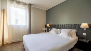 A neatly made bed in a hotel room with a large window, a chair, and two wall-mounted lamps.