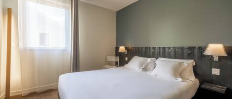 Una cama bien tendida en una habitación de hotel, con un ventanal grande, una silla y dos lámparas fijadas a la pared.