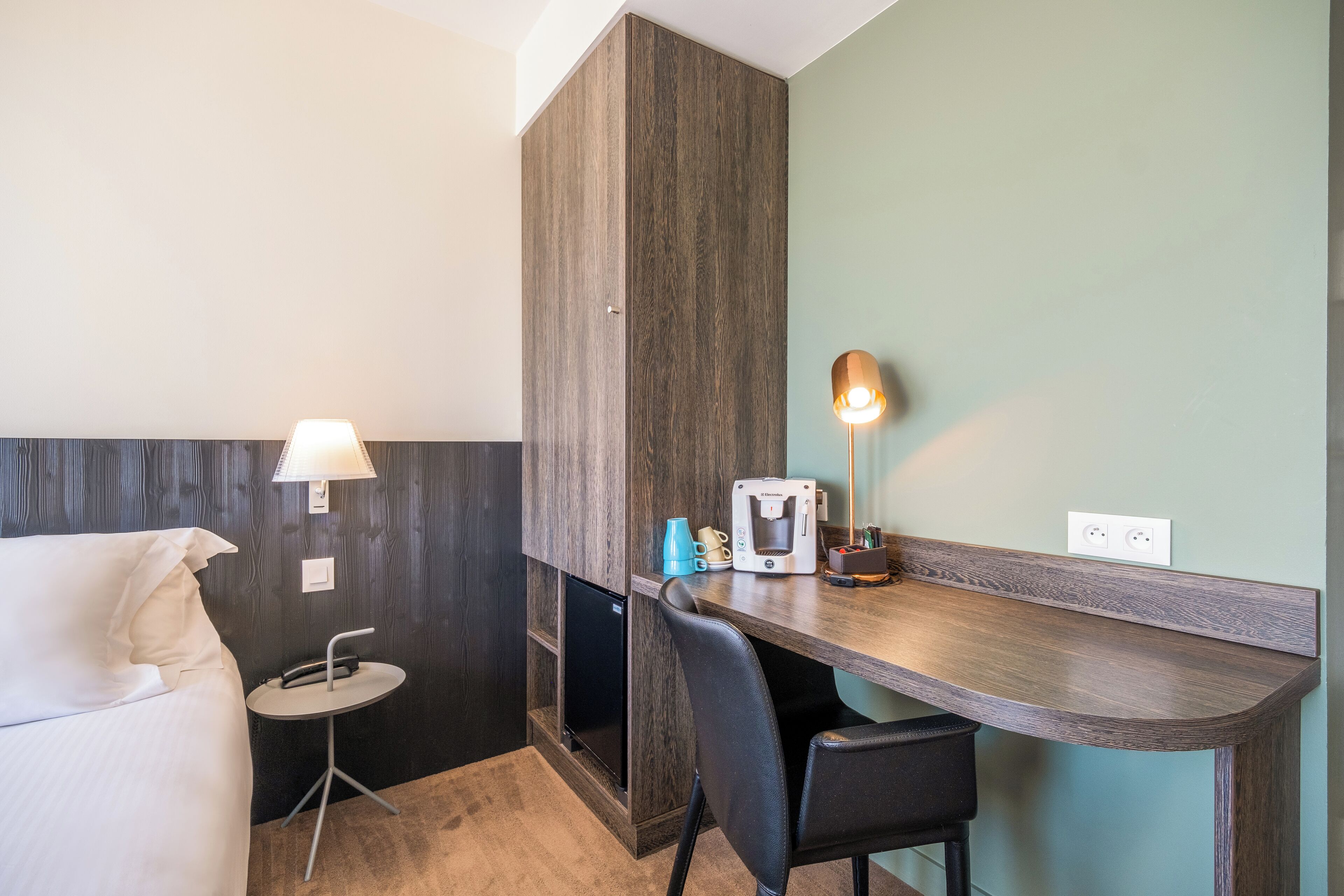 Une chambre d’hôtel de taille réduite, comprenant un lit, un bureau, une chaise, une machine à café et une lampe.