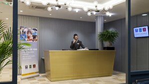 A hotel reception desk with a receptionist, a large poster advertising hotel brands, and a potted plant.