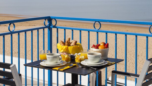 A table with breakfast items, including croissants, fruit, and coffee, on a balcony with a view of the beach and ocean.