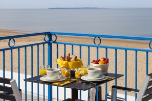 Uma mesa com itens de café da manhã, como croissants, frutas e café, numa varanda com vista para a praia e para o oceano.