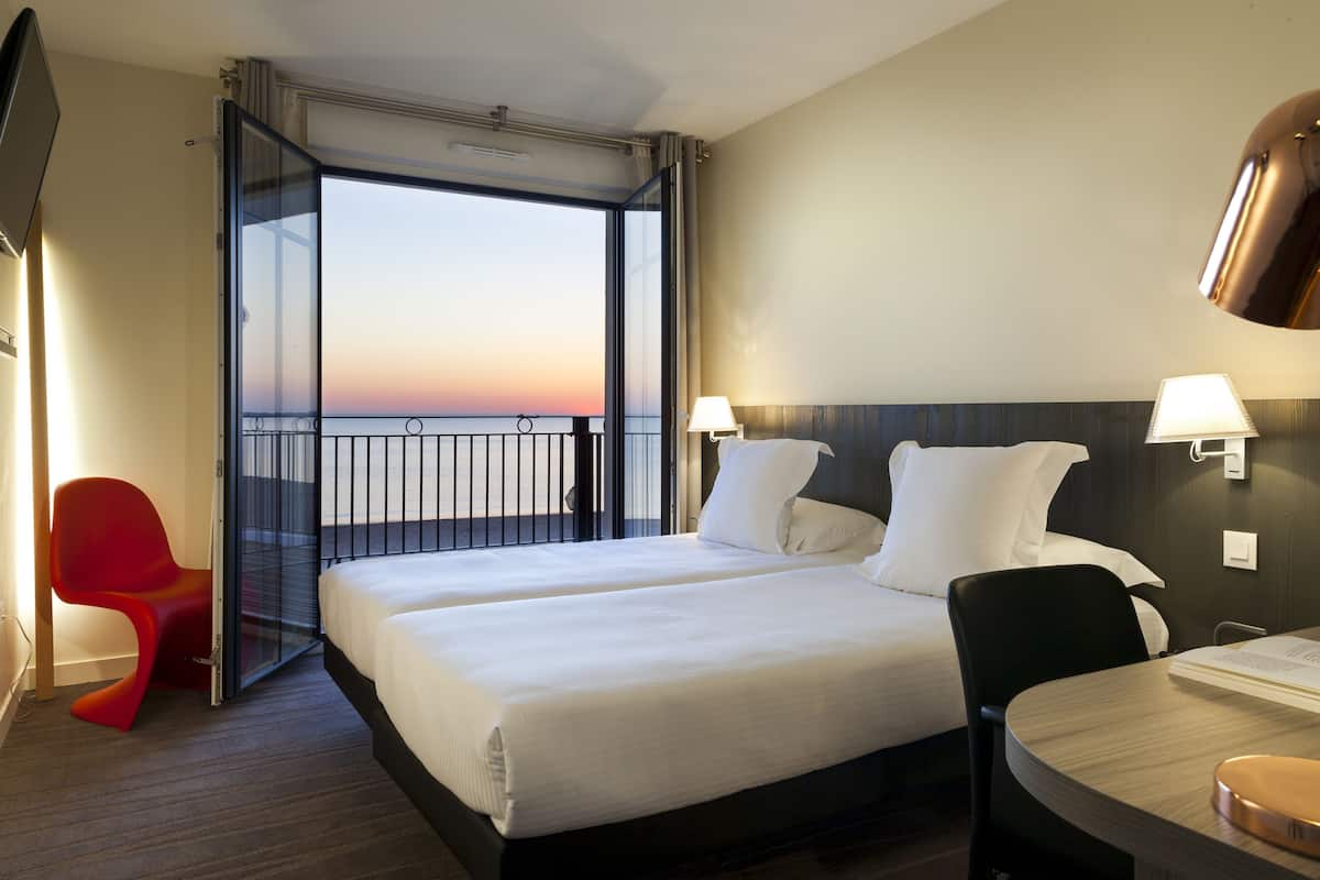Habitación de hotel con dos camas, una silla roja, una mesa y vistas al atardecer.