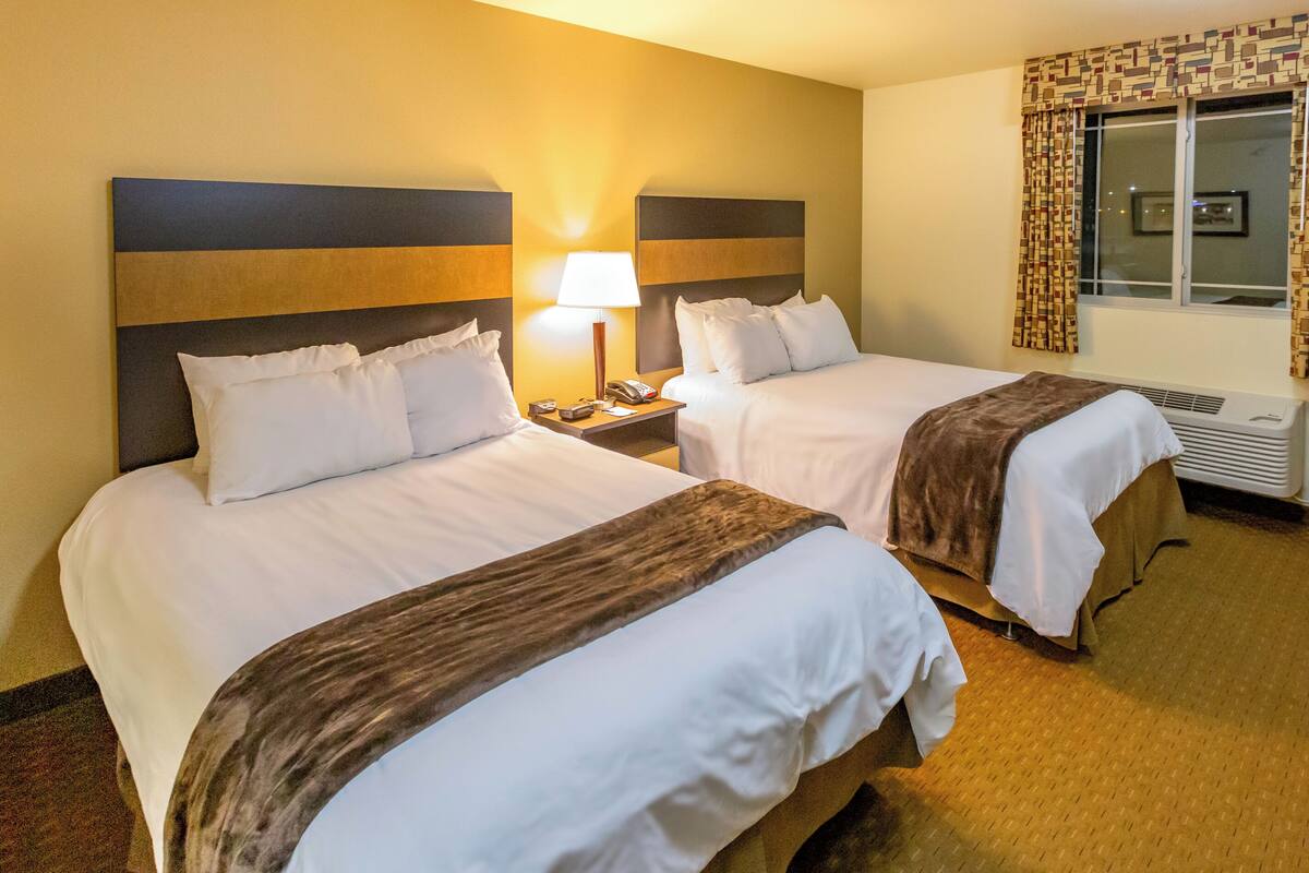A hotel room with two beds, a nightstand with a lamp, a window with curtains, and a wall-mounted air conditioner.