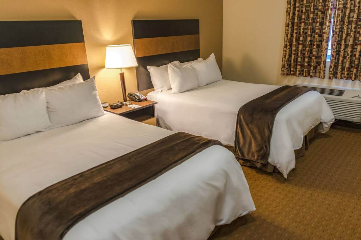 A hotel room with two beds, a nightstand with a lamp, a telephone, and an air conditioning unit.