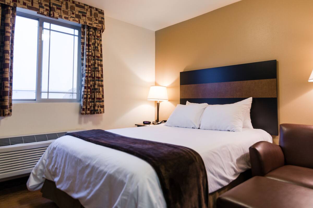 A hotel room with a bed, a chair, a nightstand, a lamp, and a window with curtains.