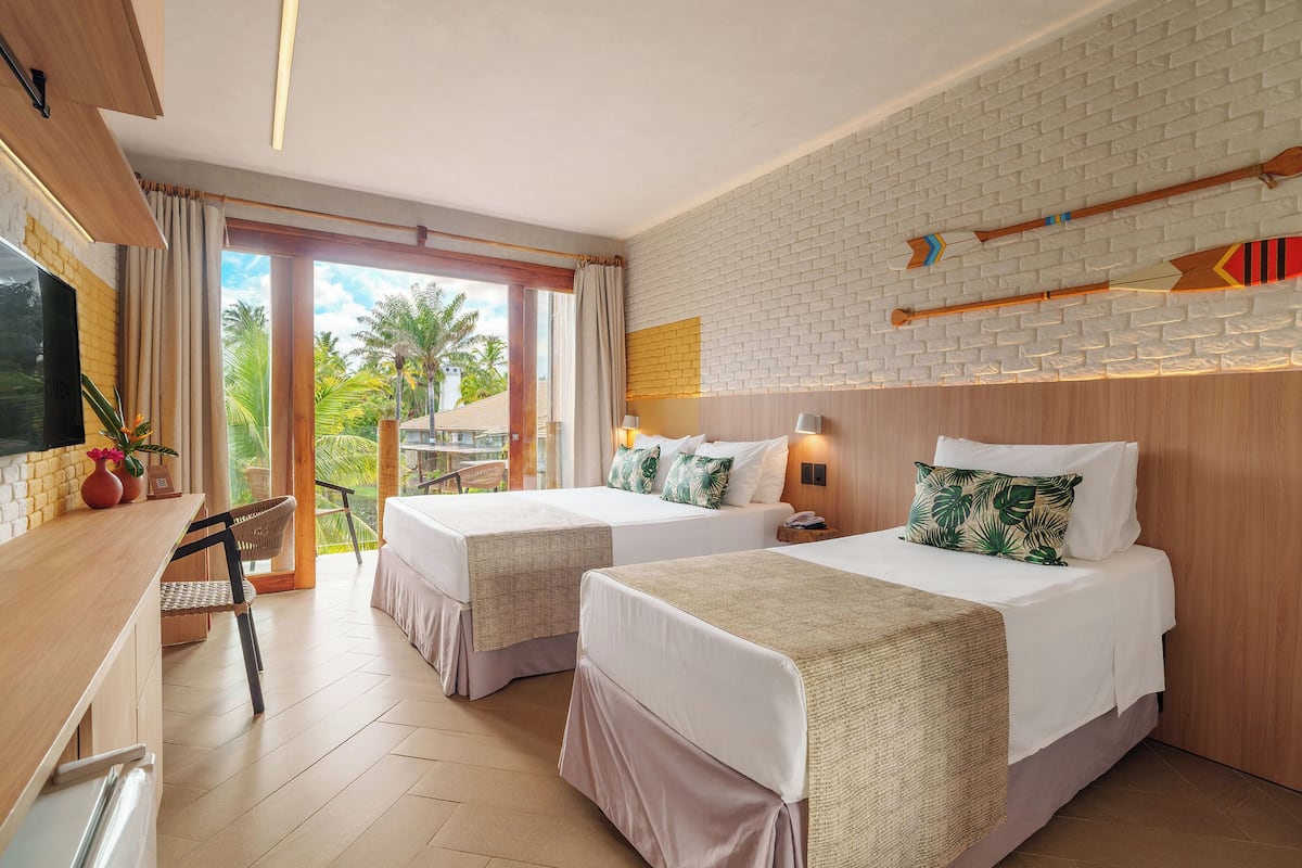 Une chambre d’hôtel avec deux lits, un bureau, une télévision et un balcon donnant sur des palmiers.
