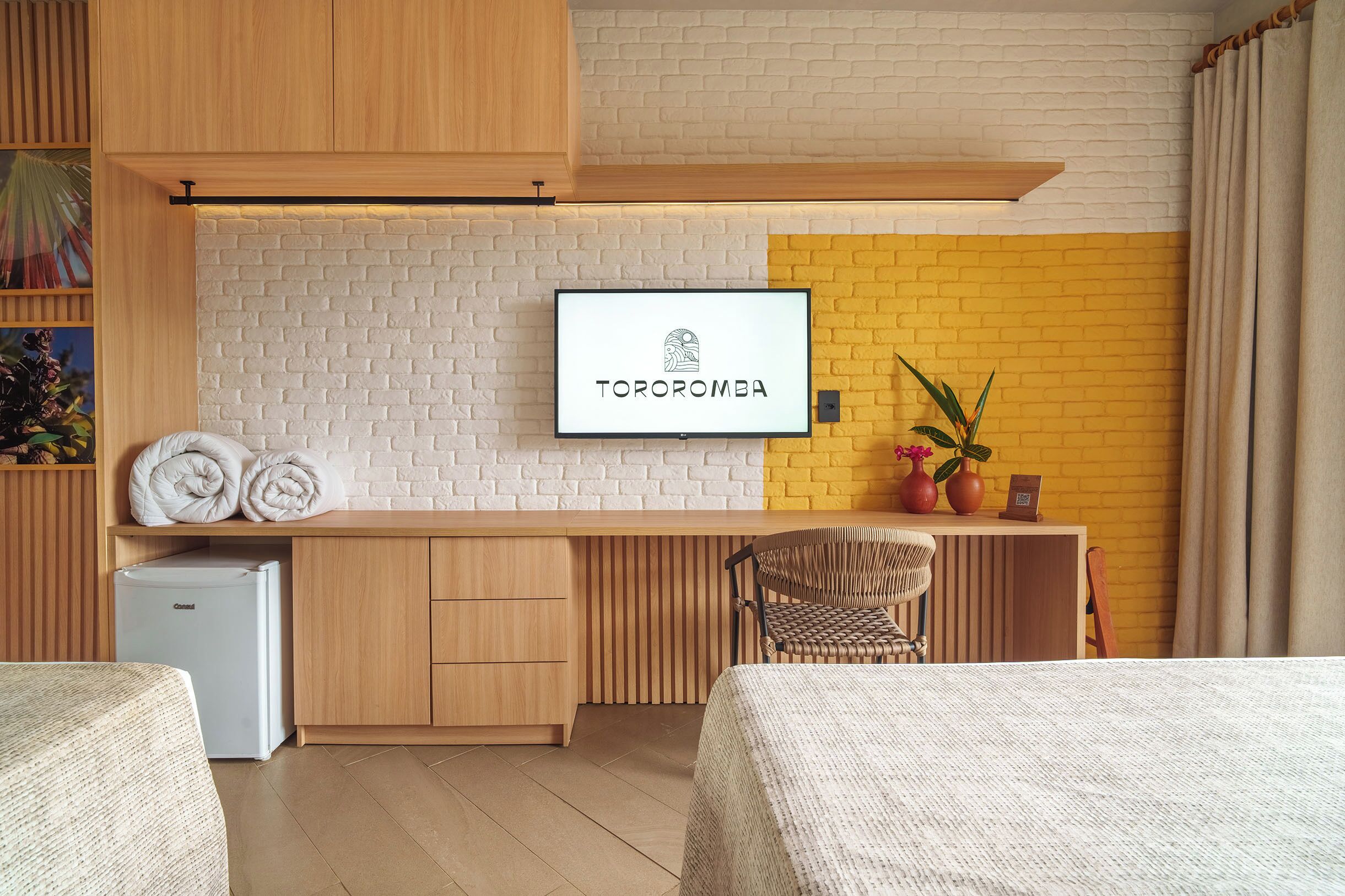 A modern hotel room with a bed, a desk, a chair, a small fridge, and a TV mounted on a brick wall.