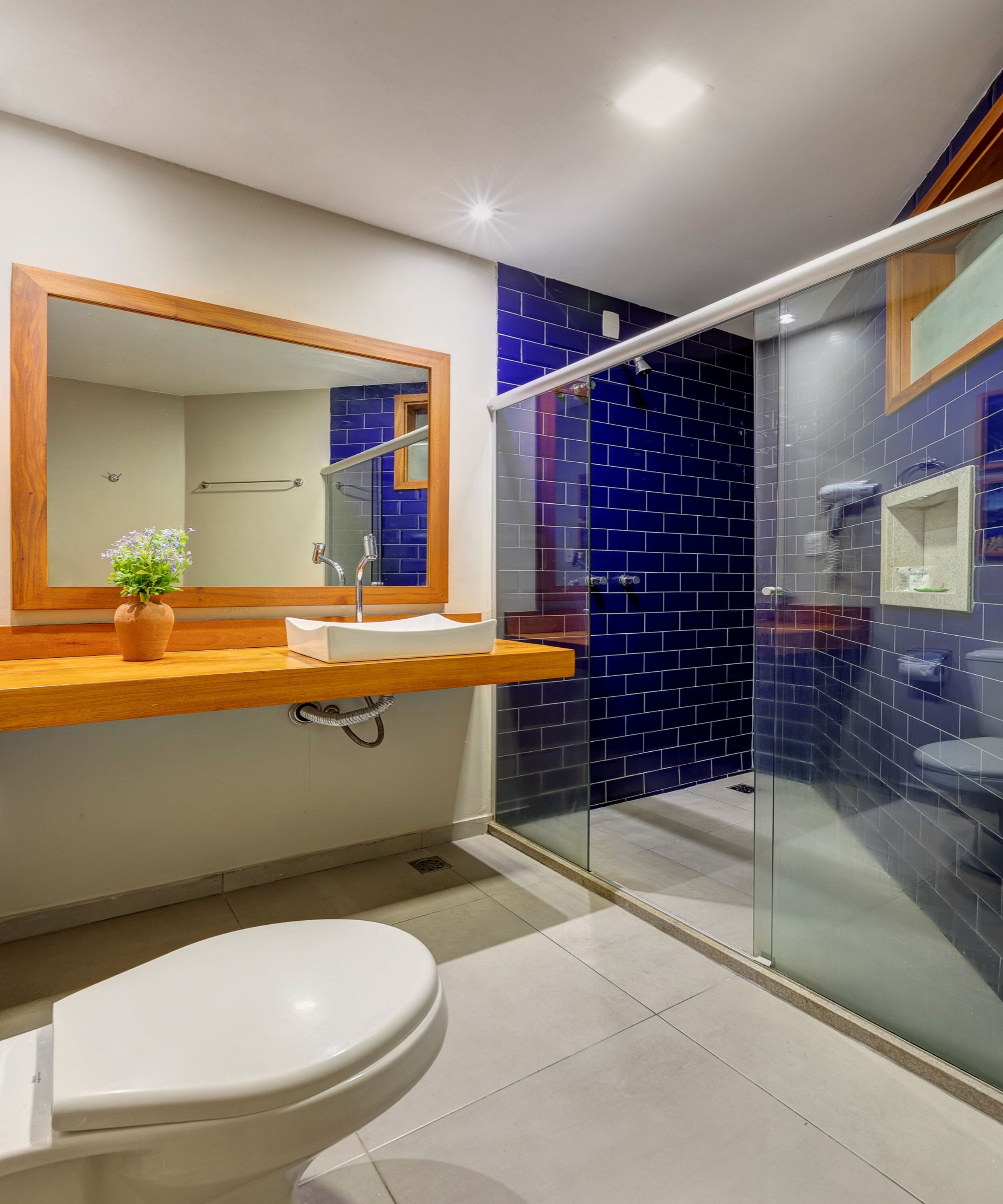 A modern bathroom with a white toilet, a wooden vanity with a mirror, and a glass-enclosed shower area with blue tiles.