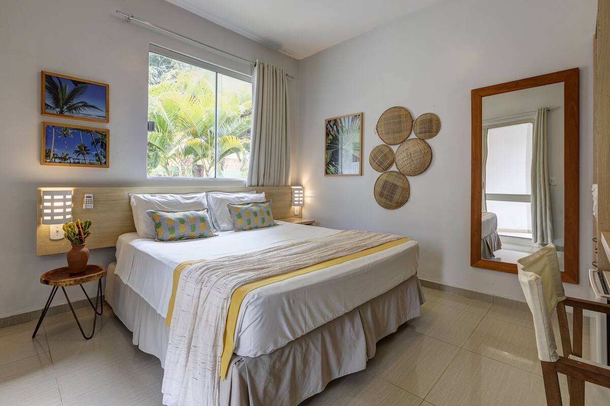 Une chambre spacieuse avec un grand lit, une table de chevet avec une lampe, une plante en pot et une vue sur les palmiers par la fenêtre.