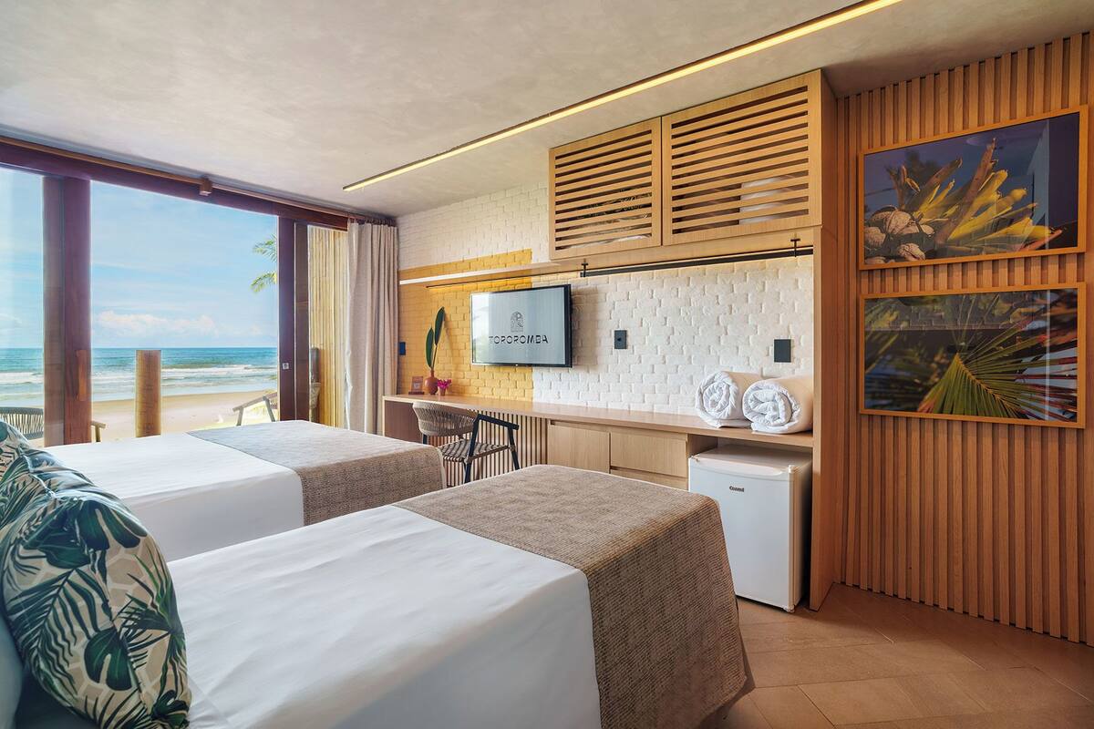 Une chambre d’hôtel avec deux lits, une télévision, un petit réfrigérateur, un panneau mural en bois et une vue sur la plage.