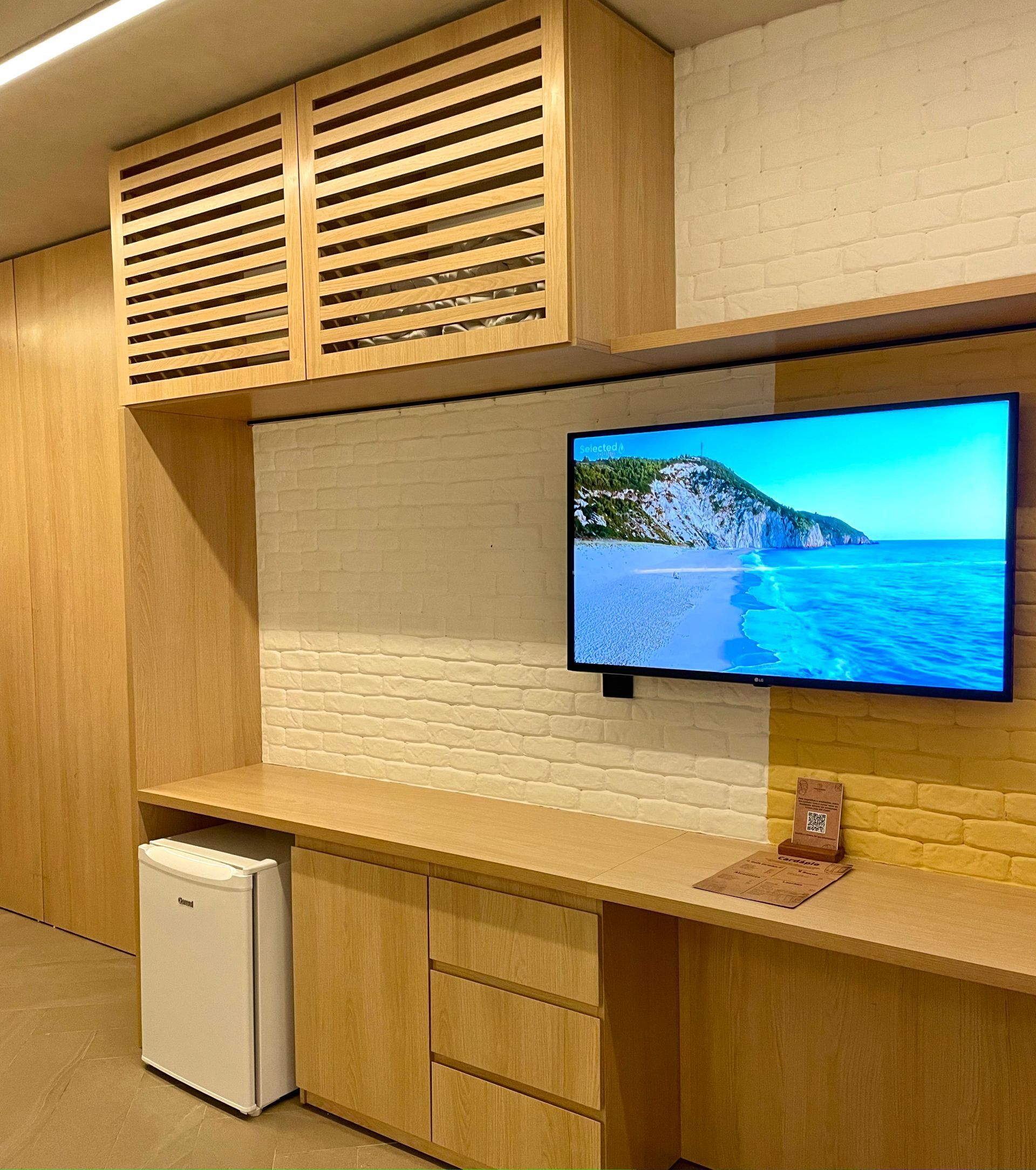 A modern kitchen with wooden cabinets, a built-in refrigerator, and a flat-screen TV mounted on a brick wall.