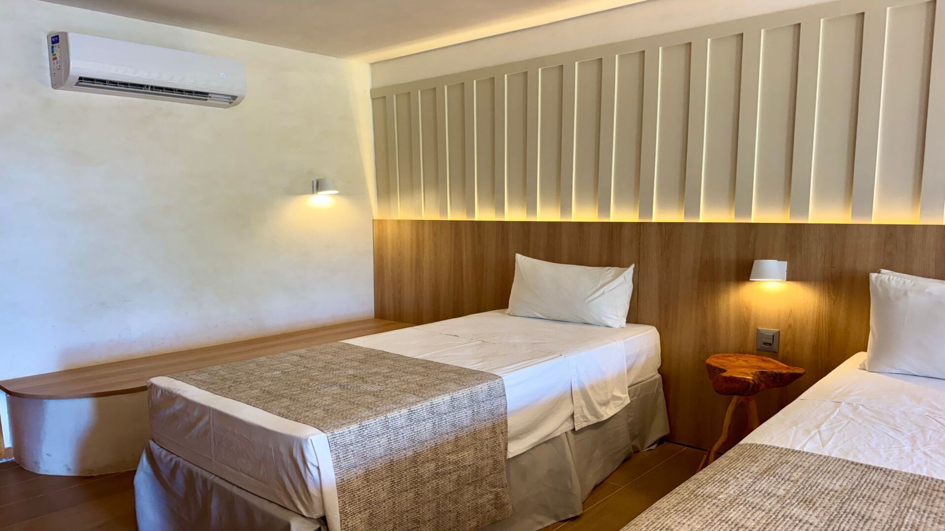 A hotel room with a bed, bedside tables, a nightstand, an air conditioning unit, and a wall-mounted light fixture.