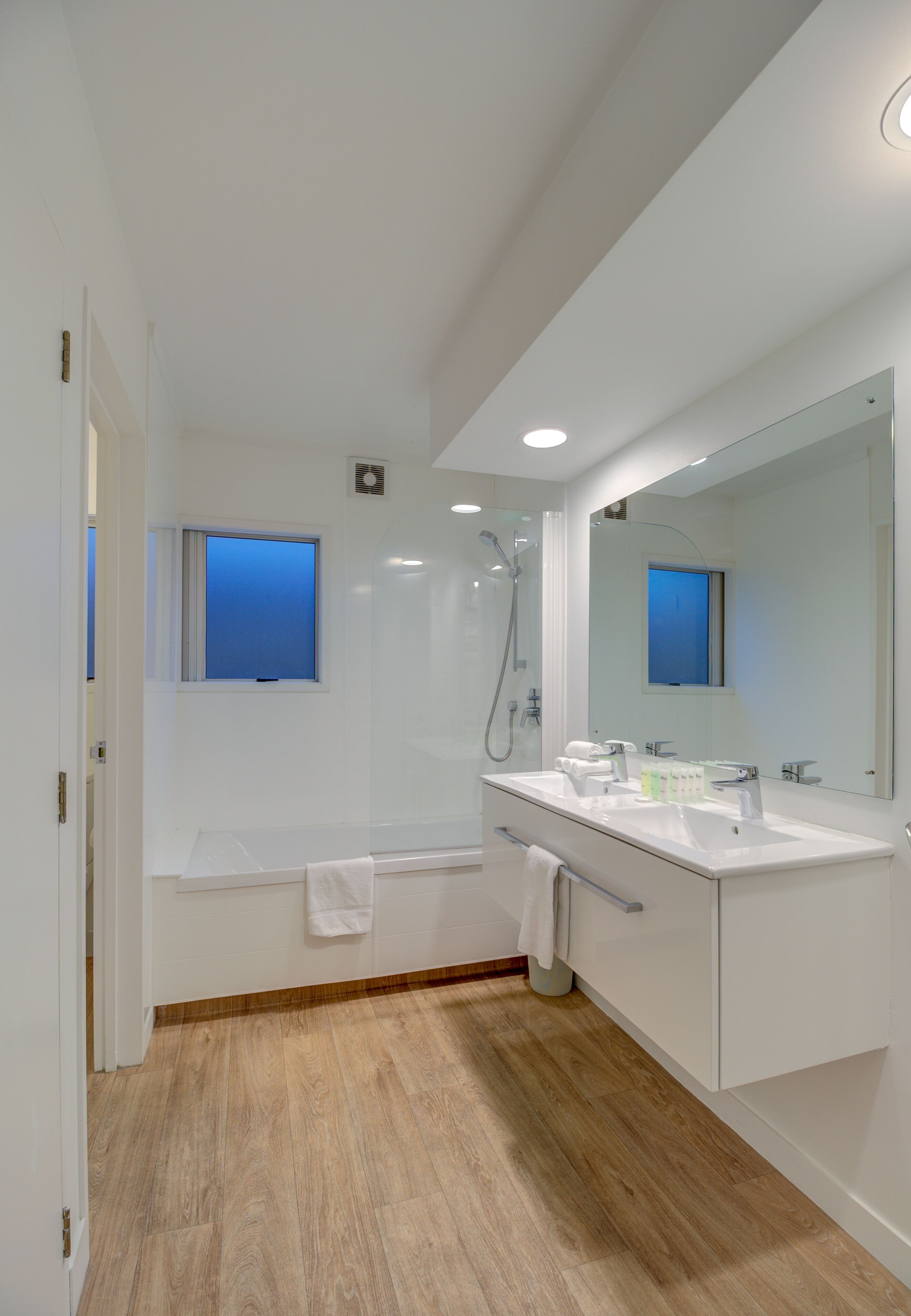 A modern bathroom with a large mirror, two sinks, a shower, and a window.