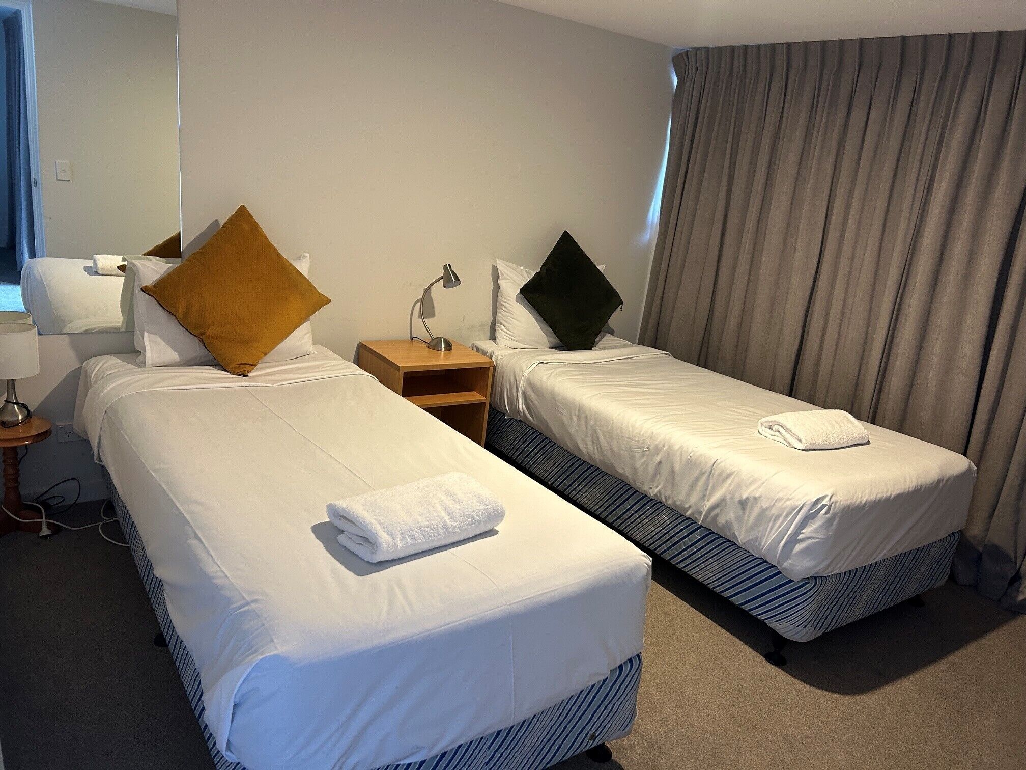 A hotel room with two beds, a nightstand, a lamp, and a window with curtains.