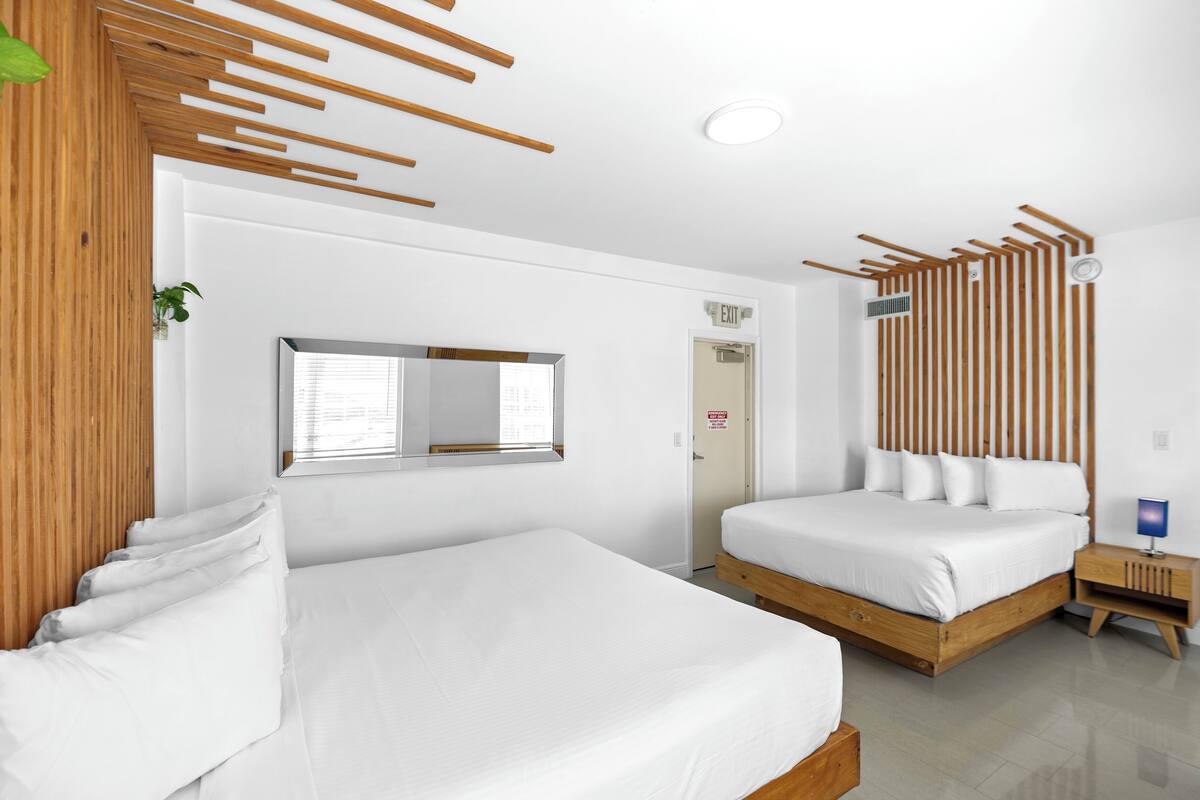 A modern hotel room with two single beds, a wooden headboard, a small bedside table with a lamp, and a mirror on the wall.