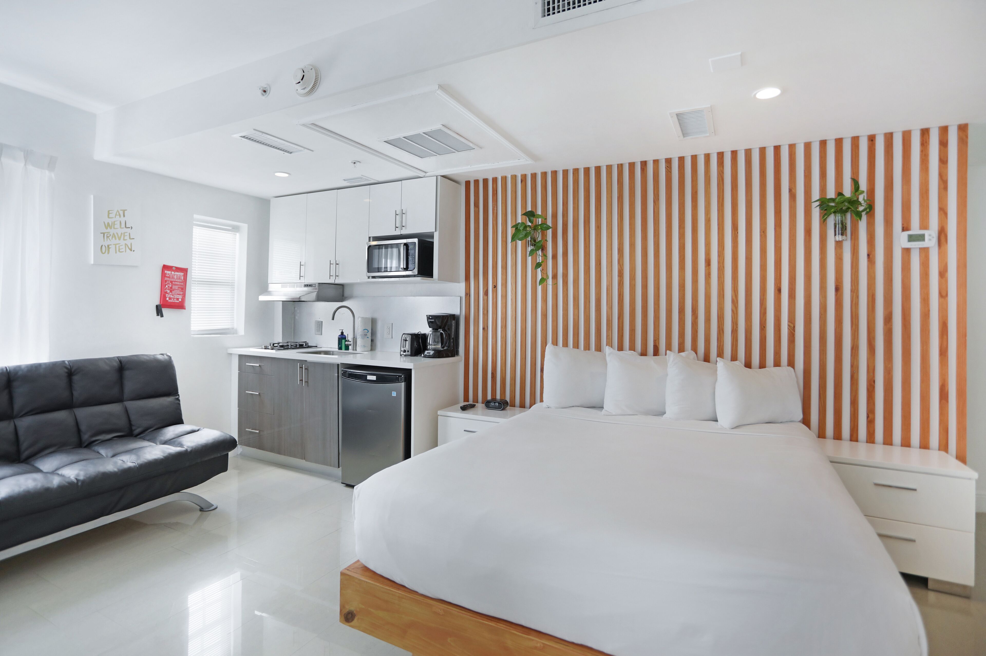 A modern hotel room with a large bed, a sofa, a kitchenette, and a striped wall.