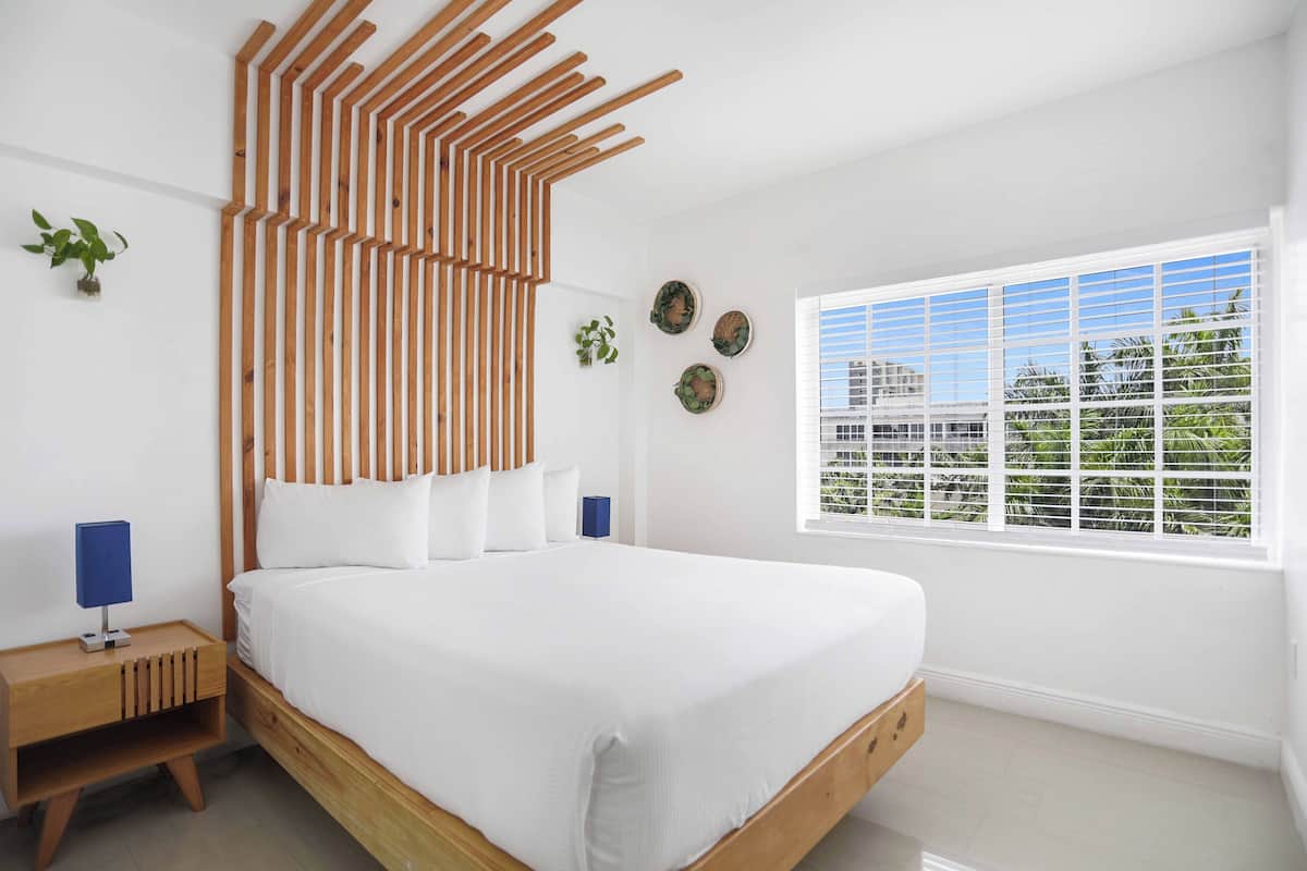 A modern bedroom with a wooden headboard, a large bed, a nightstand with a lamp, and a window with blinds.