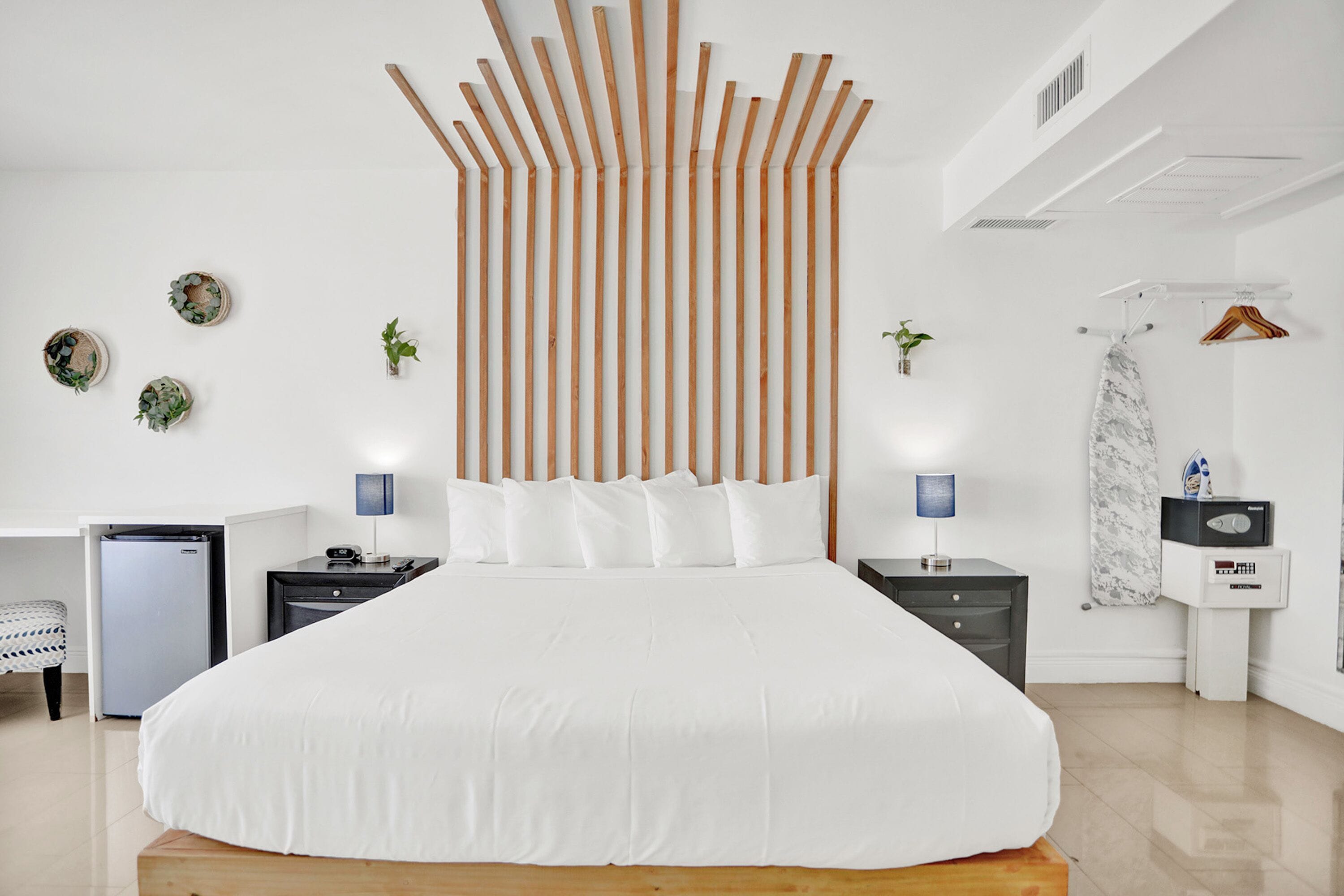 A modern bedroom with a large bed, wooden slats on the wall, bedside tables, a small refrigerator, and a hanging clothes rack.