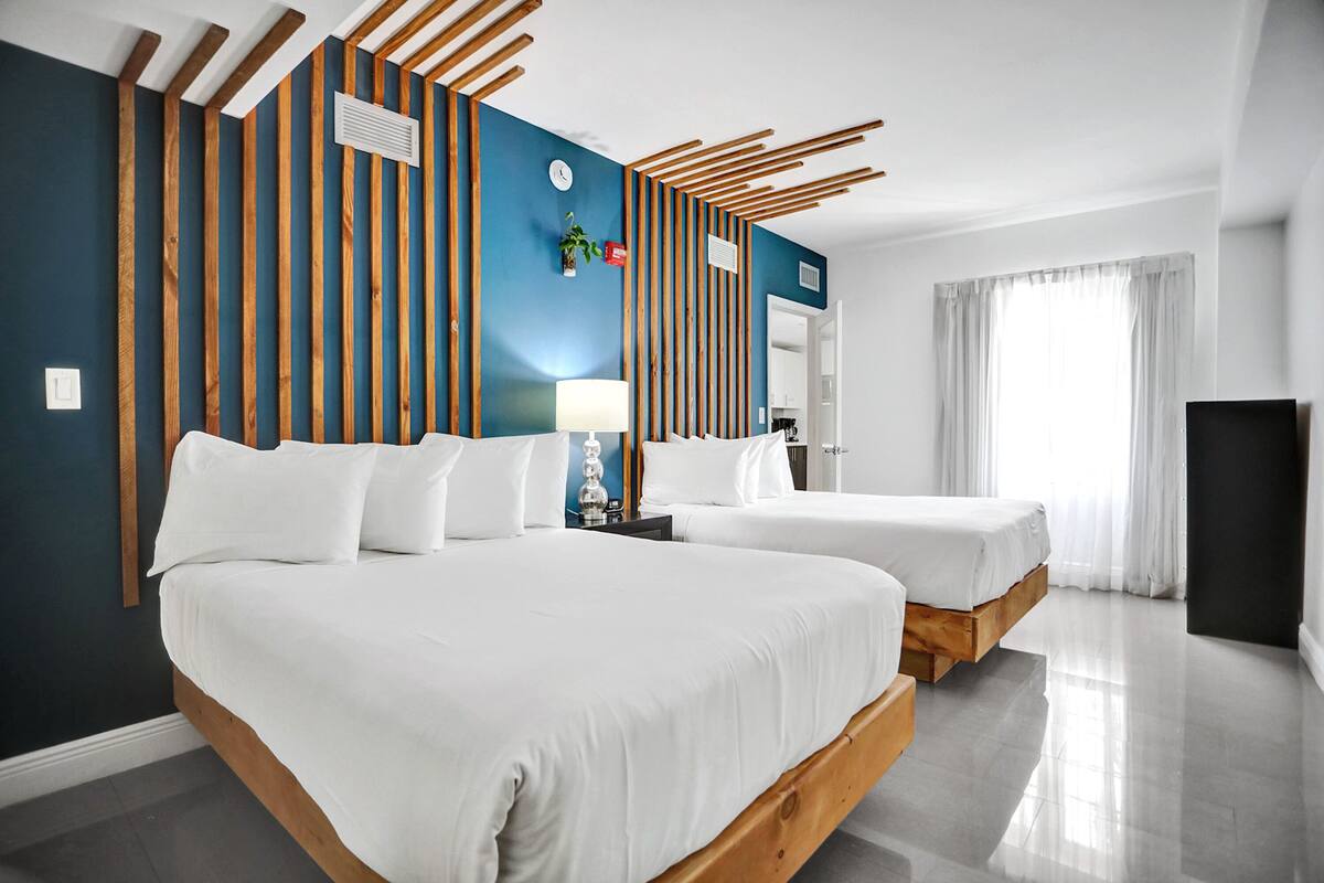 A modern hotel room with two beds, a large window with sheer curtains, and a wooden ceiling design.