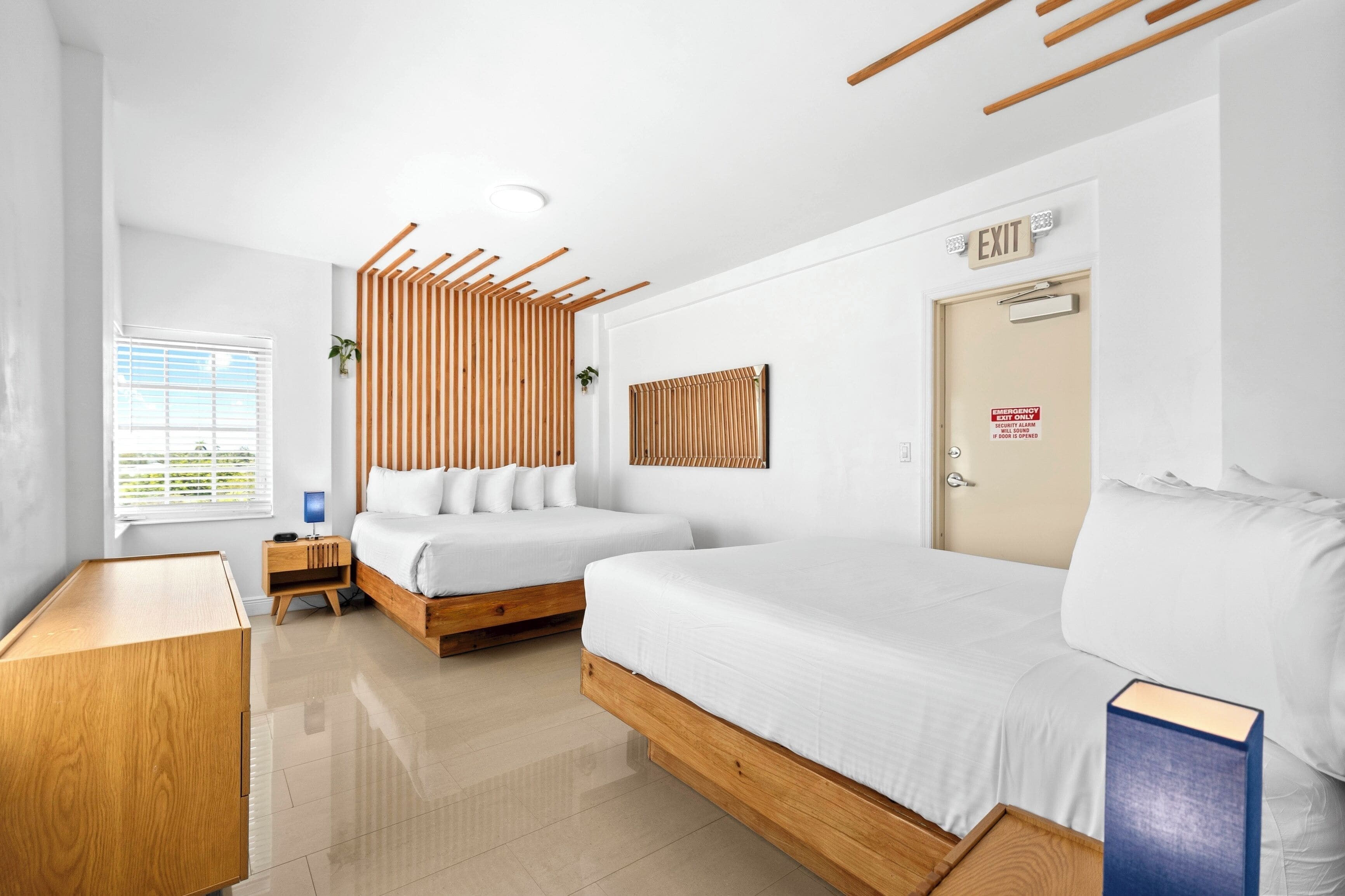 A modern hotel room with two beds, a wooden headboard, a bedside table, a nightstand, a window with blinds, and an exit door.