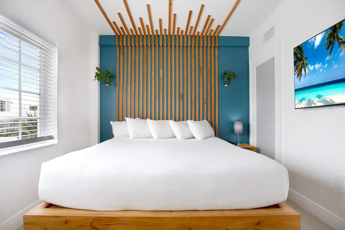 A bedroom with a large bed, a wooden headboard, a view of the city, and a tropical beach scene on the wall.