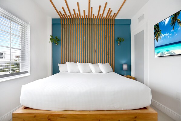 A bedroom with a large bed, a wooden headboard, a view of the city, and a tropical beach scene on the wall. - Hotel Belleza (Miami Beach)