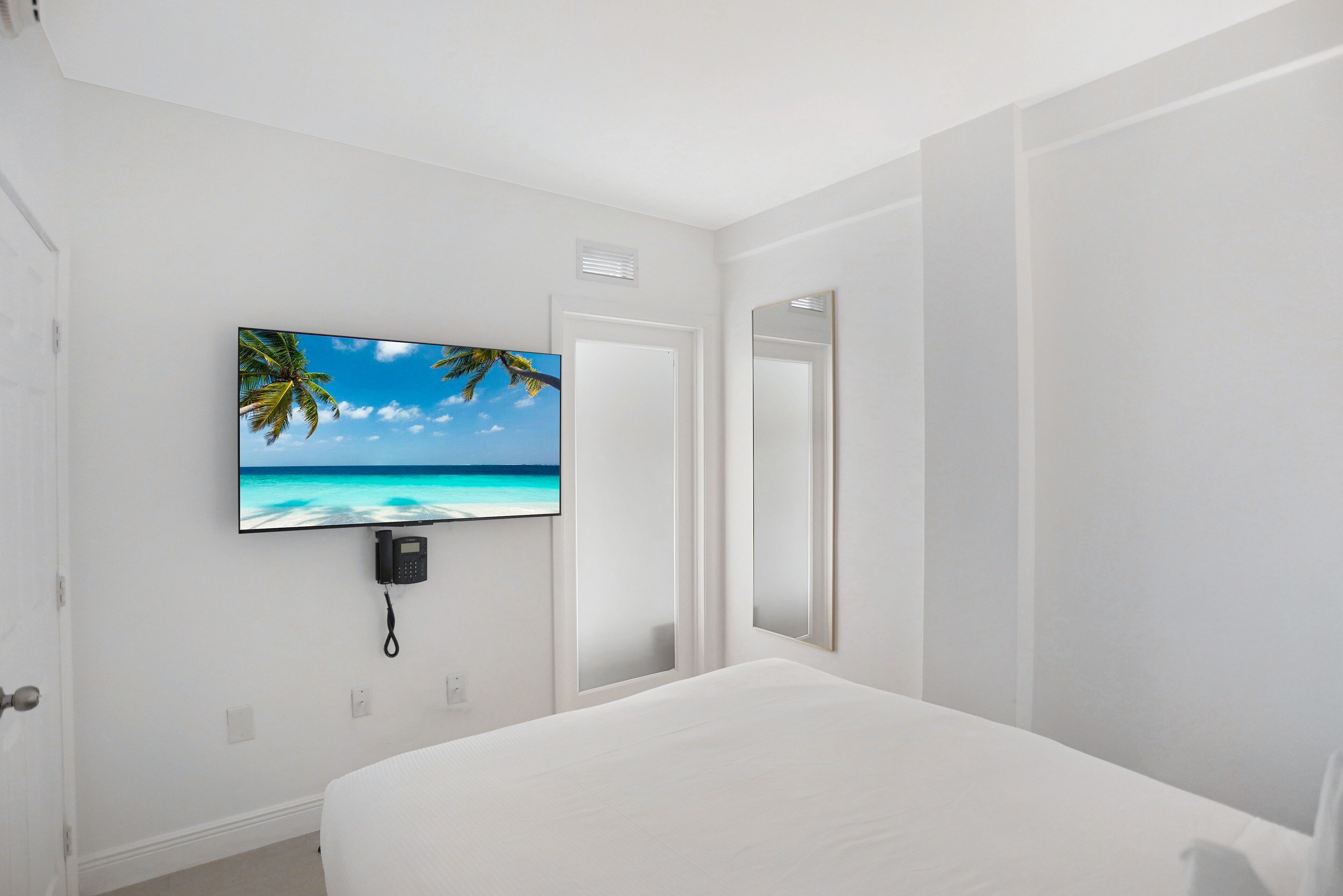 A hotel room with a bed, a mounted TV displaying a beach scene, a phone, and a door.
