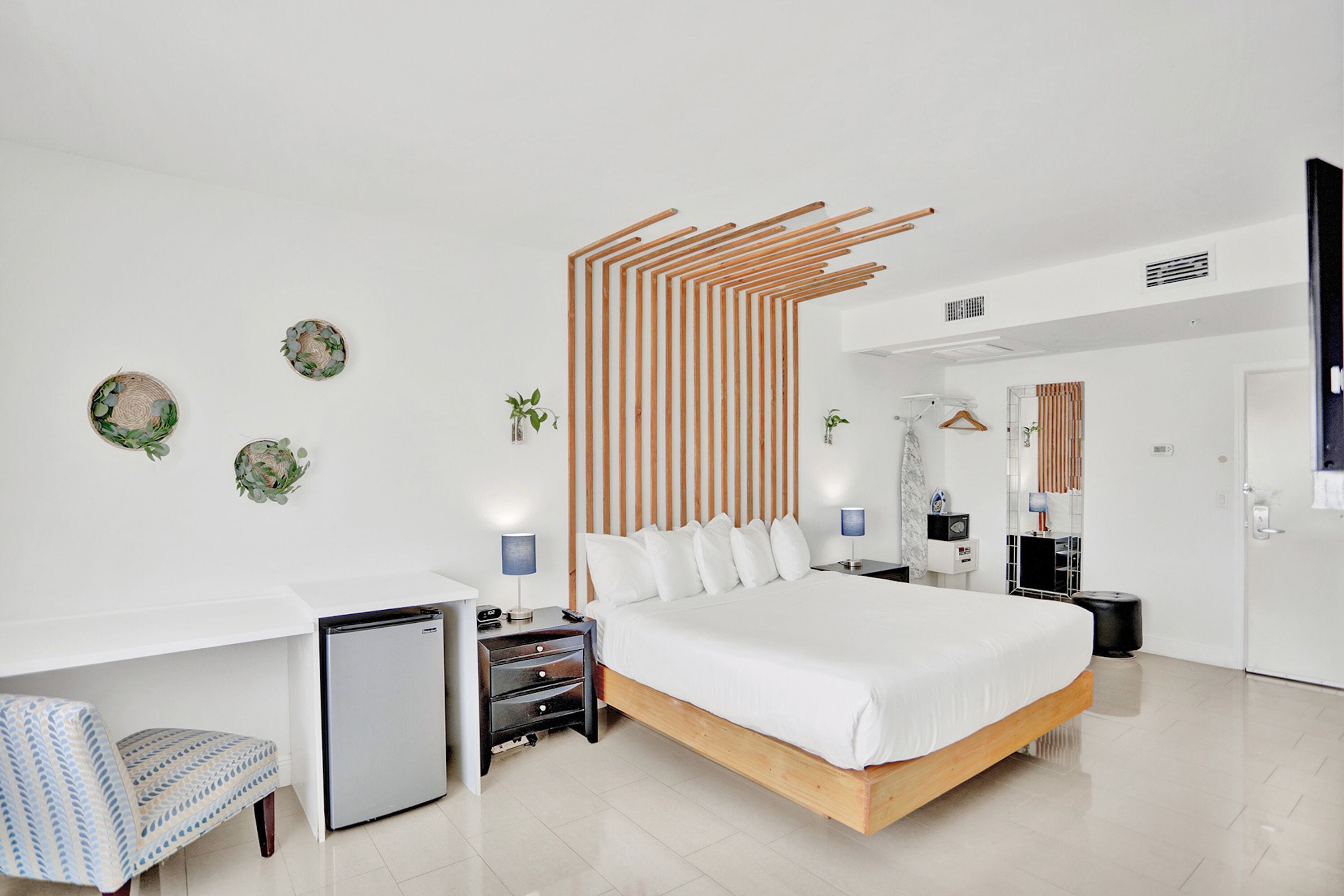 A modern hotel room with a large bed, a desk, a chair, a small fridge, and decorative wall art.