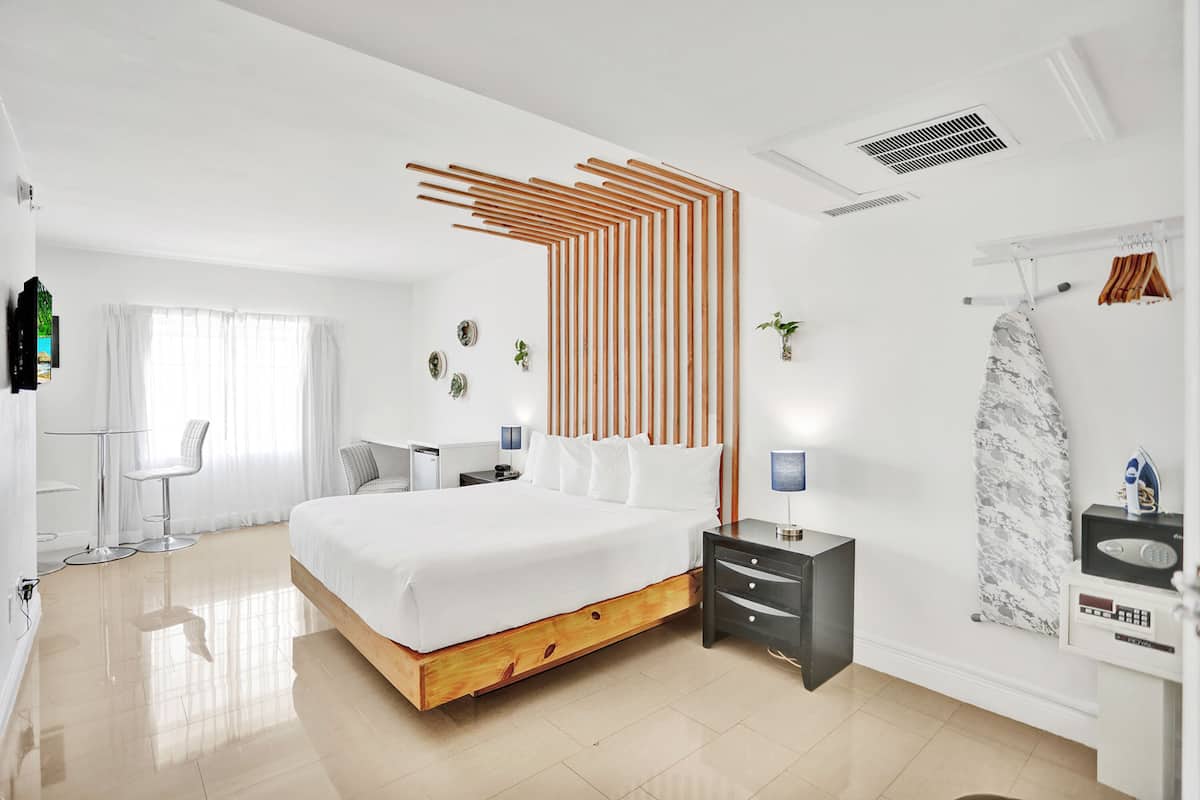 A modern hotel room with a large bed, a bedside table, a chair, a desk, and a bathroom with a shower and a towel rack.