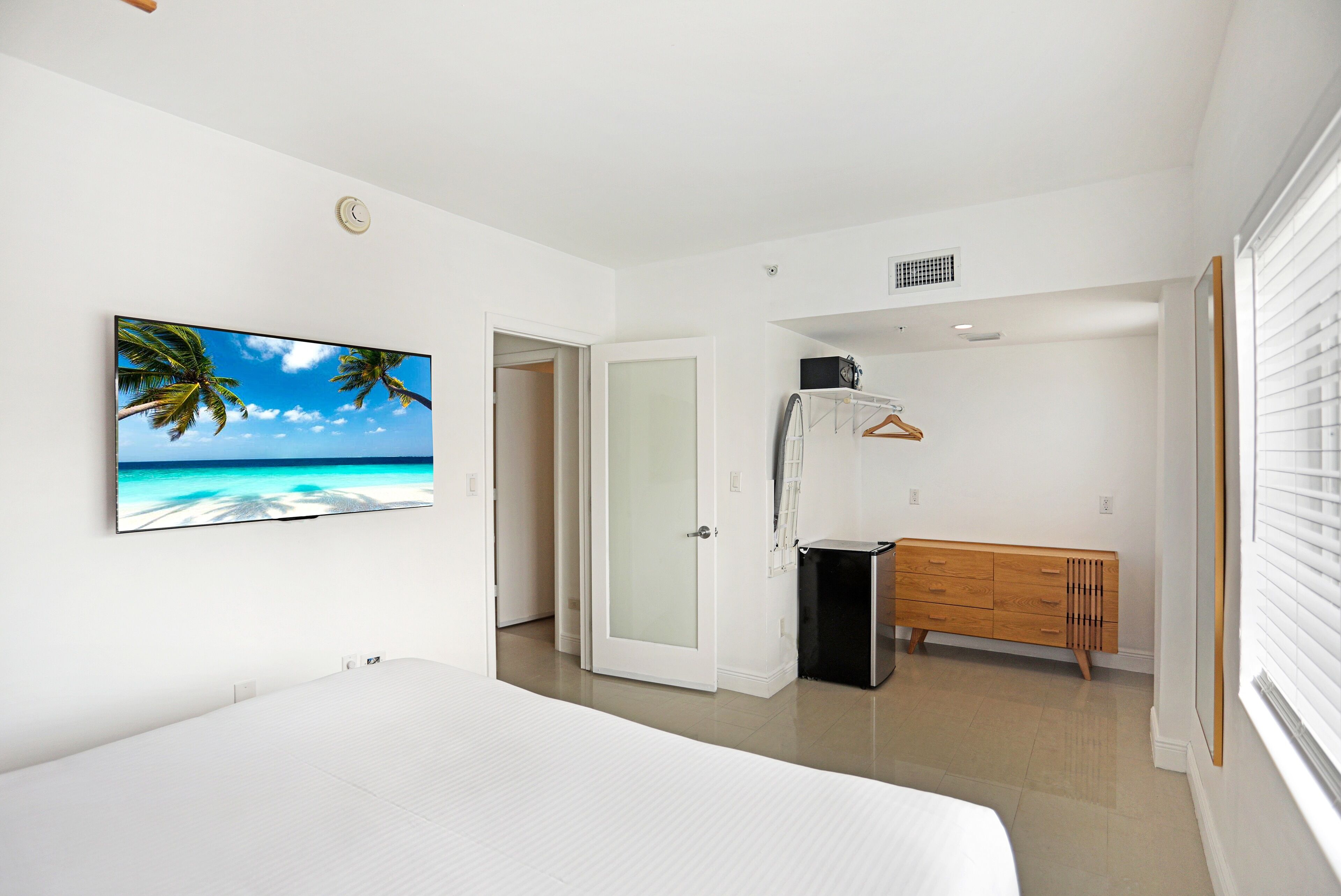 A bedroom with a bed, a wardrobe, a bench, a door, and a wall-mounted TV displaying a beach scene.