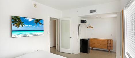 A bedroom with a bed, a wardrobe, a bench, a door, and a wall-mounted TV displaying a beach scene.