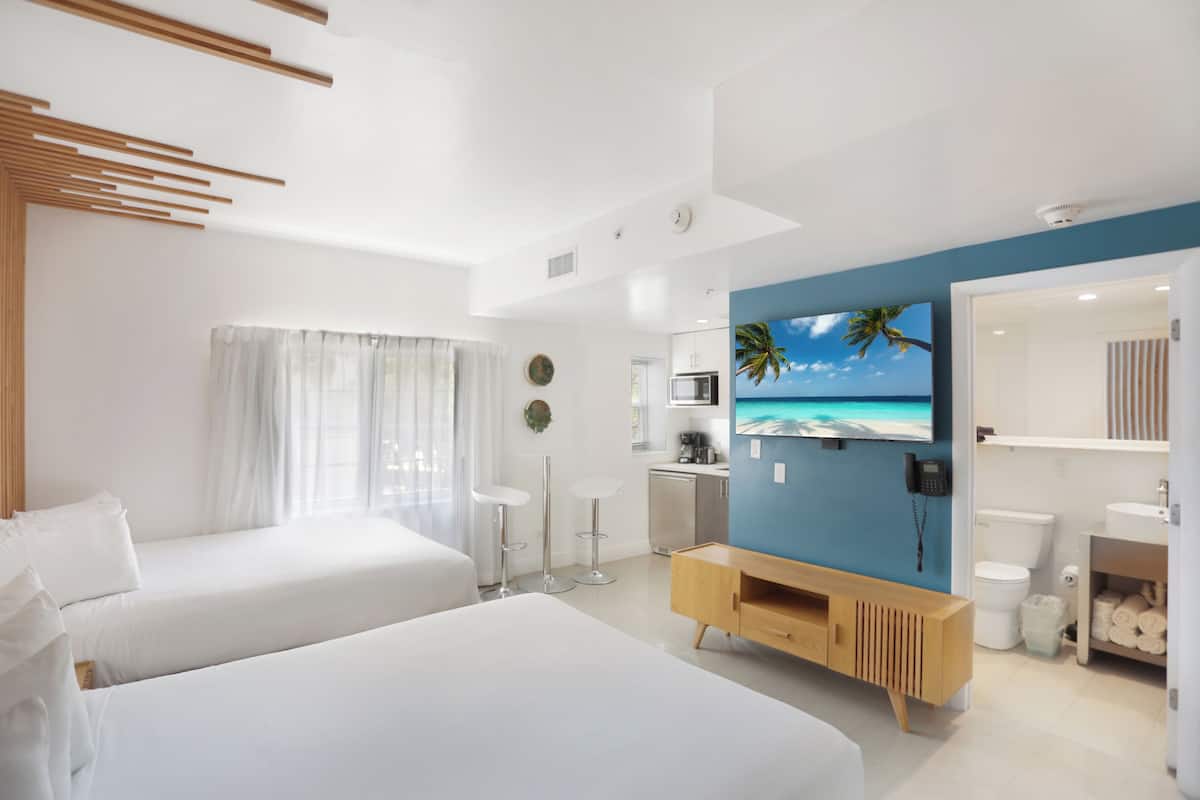 A modern hotel room with two beds, a kitchenette, a bathroom, and a large TV screen displaying a beach scene.