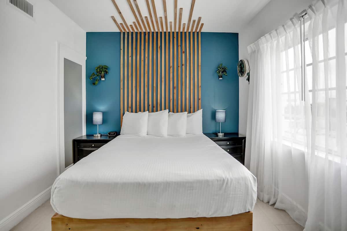 A modern bedroom with a large bed, wooden headboard, two bedside tables with lamps, and a view of the outdoors through sheer curtains.