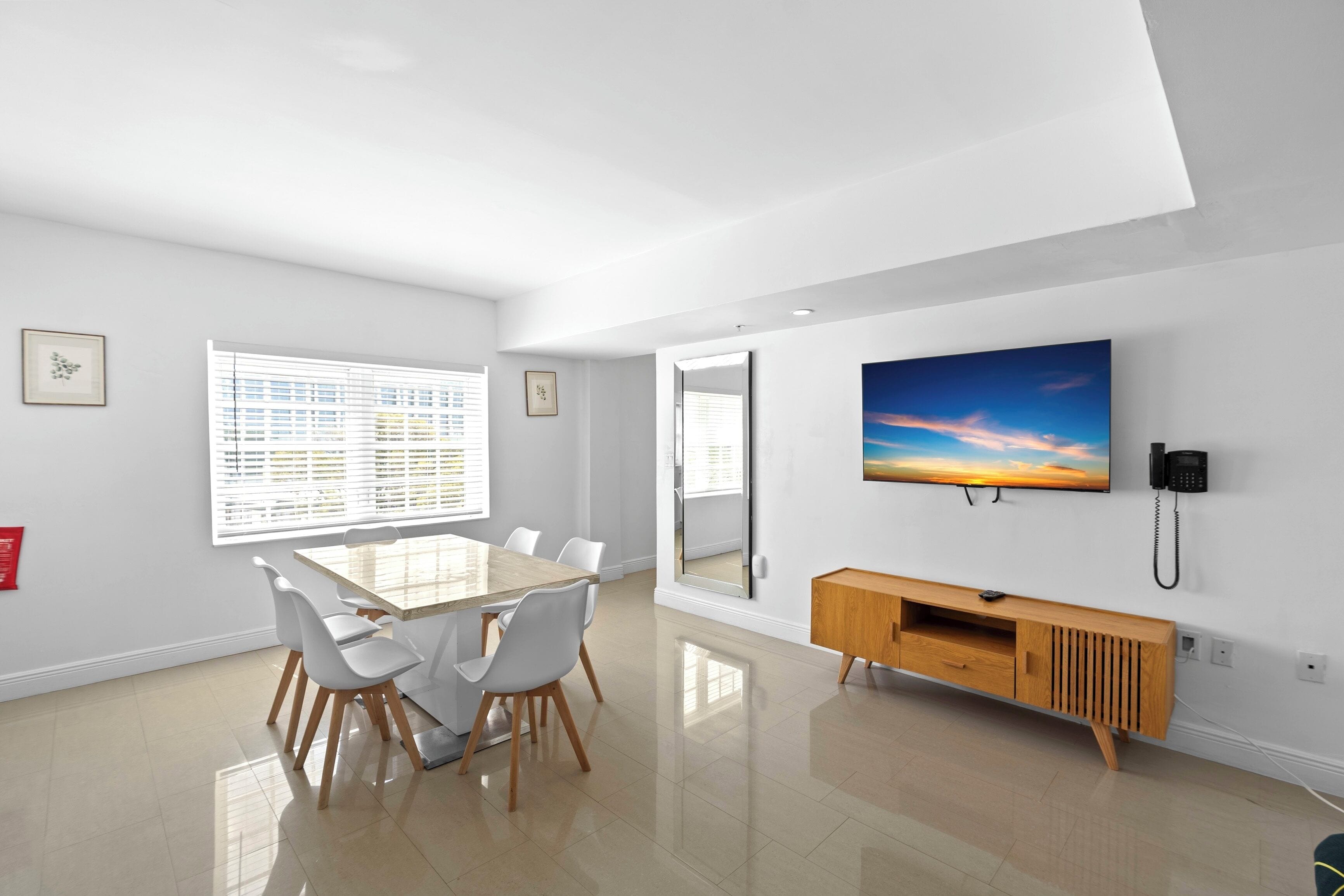 A modern dining area with a large table, white chairs, a wooden cabinet, and a mounted TV.