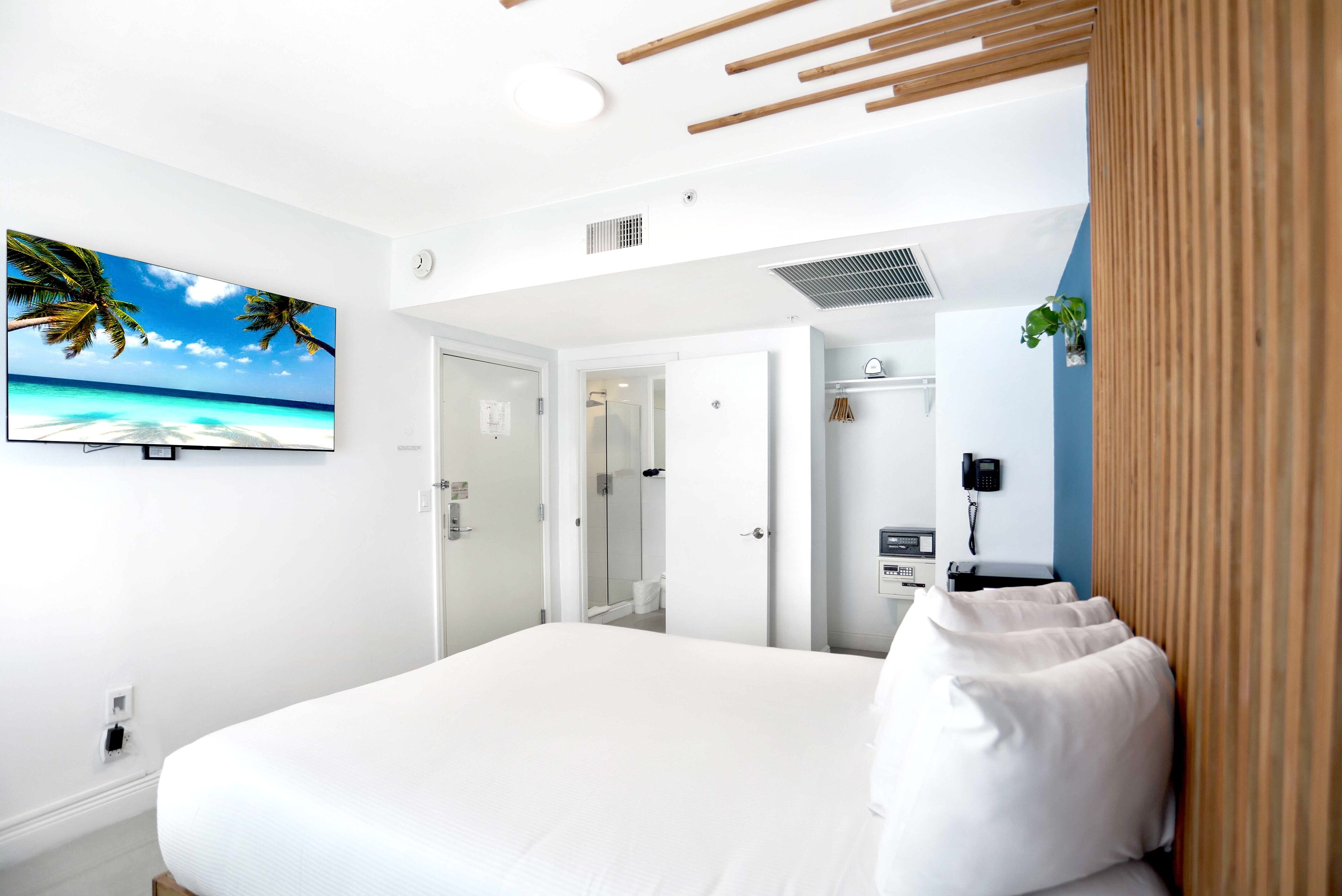A hotel room with a large bed, a television displaying a beach scene, a bathroom with a shower, and a phone on the wall.