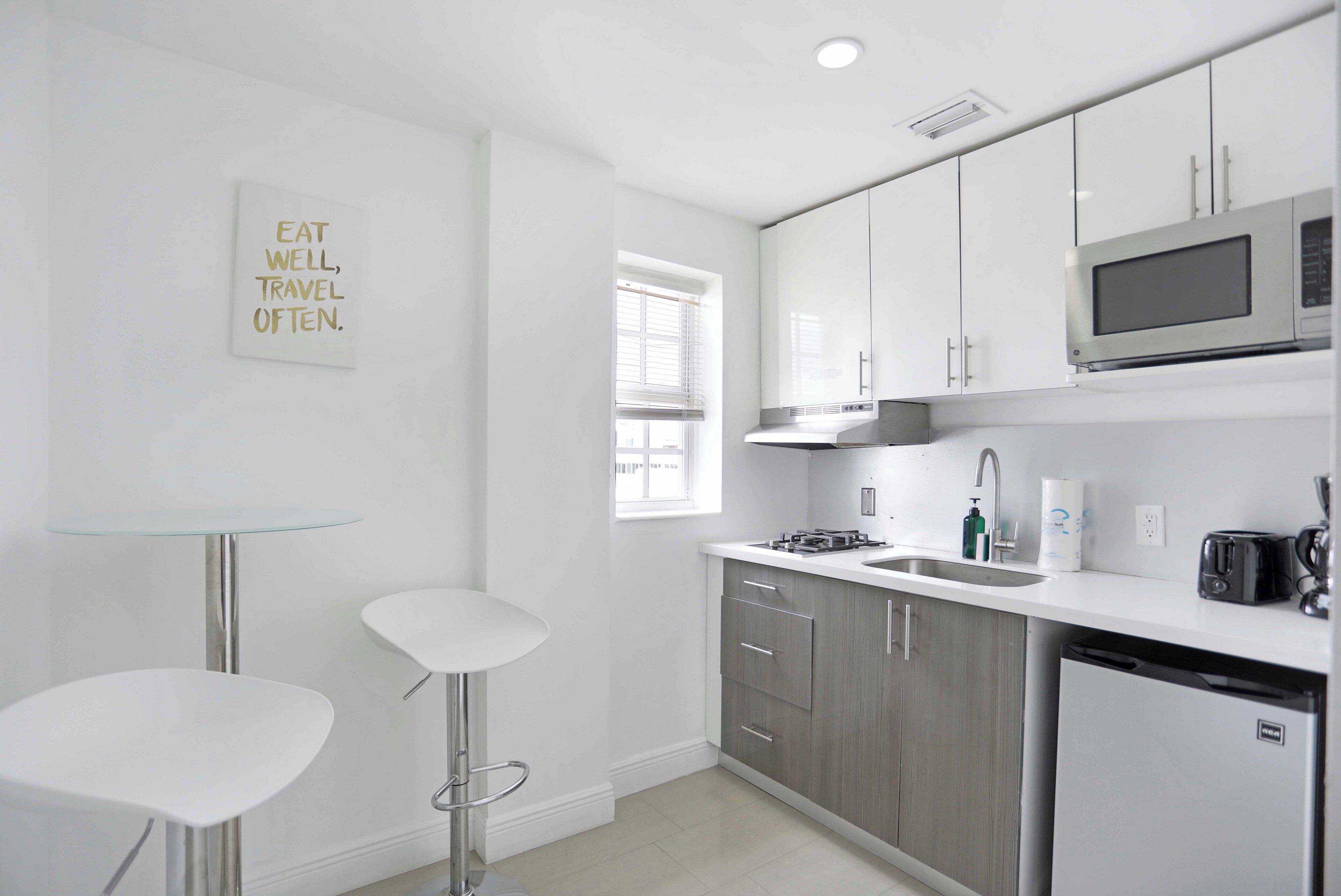 A modern kitchen with white cabinets, a stainless steel microwave, a sink, and a glass table with two bar stools.