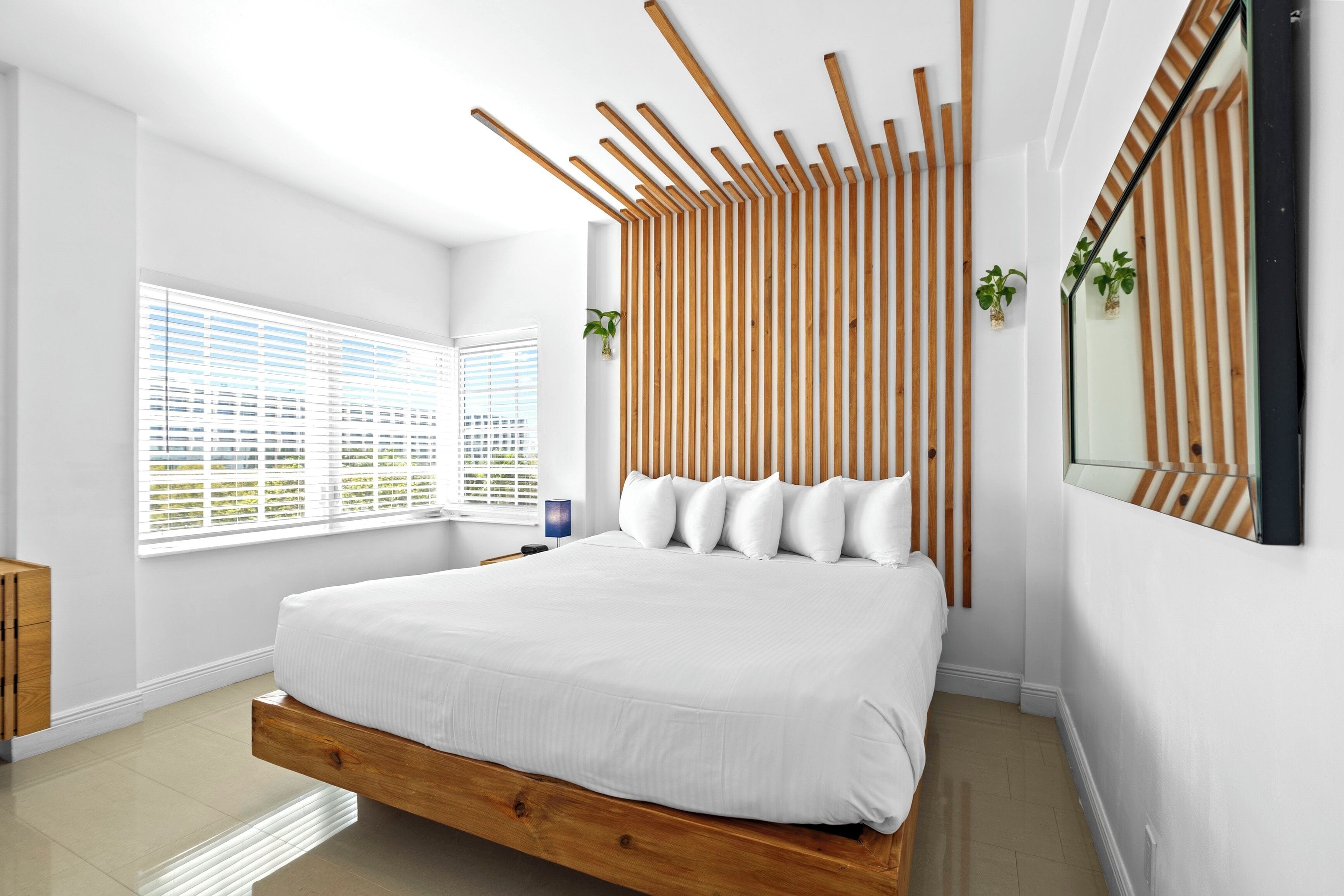 A modern bedroom with a wooden bed frame, white bedding, a slatted wooden headboard, and a large window with blinds.