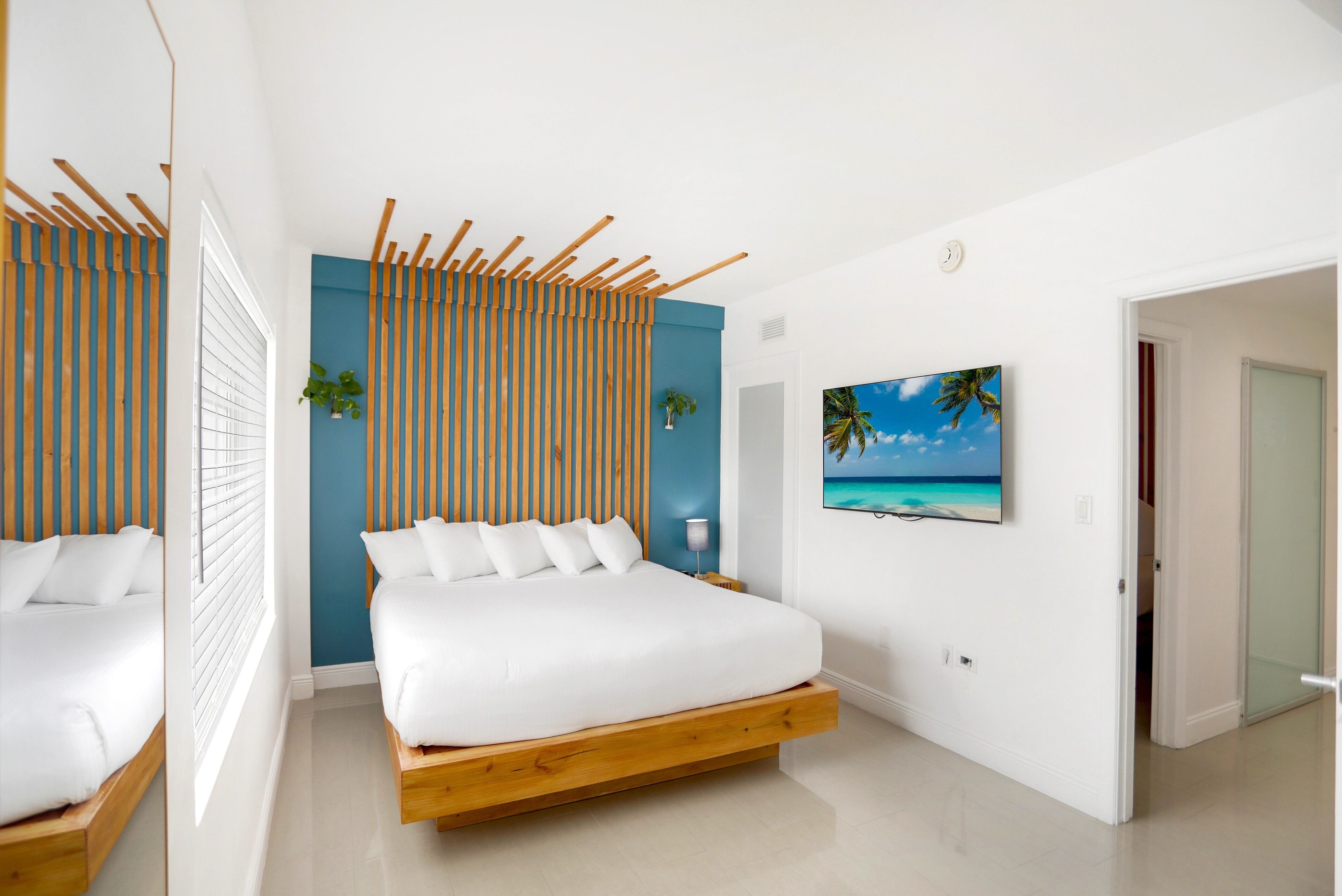 A modern bedroom with a wooden bed frame, white bedding, and a tropical-themed painting on the wall.