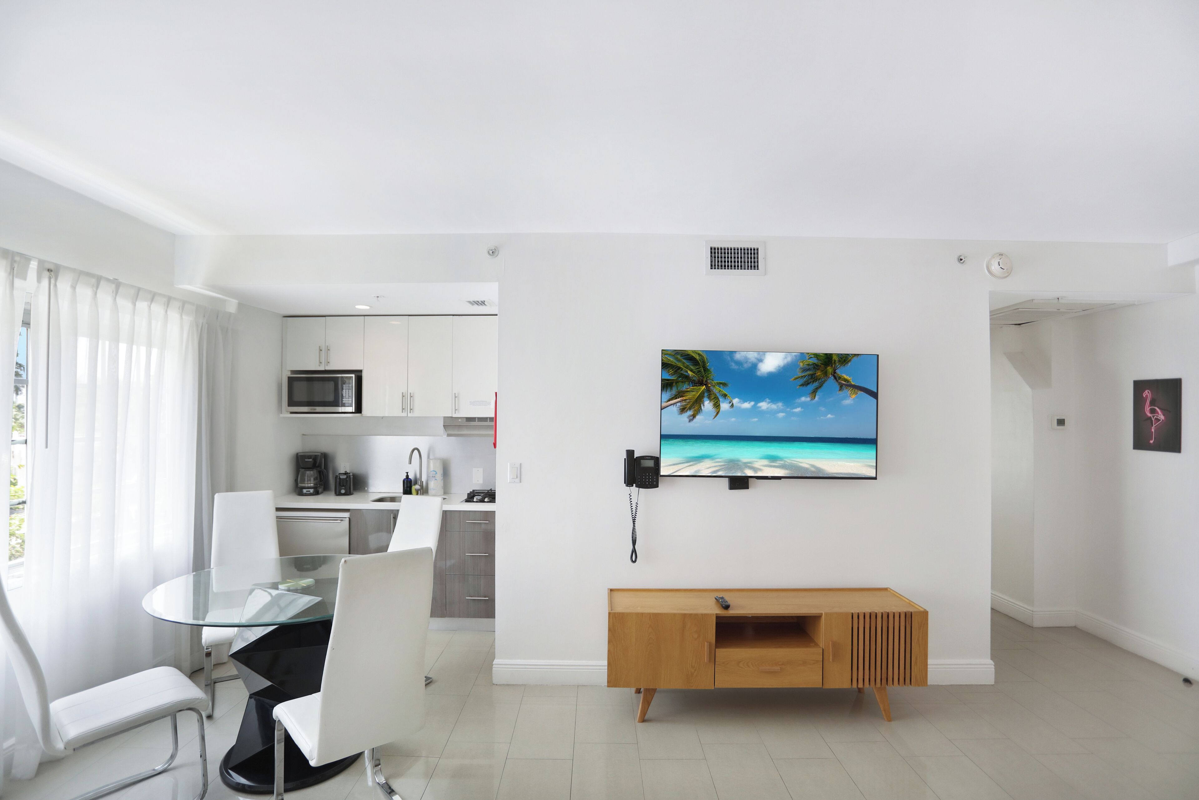 A modern living room with a glass dining table, white chairs, a wooden console table, and a mounted TV displaying a beach scene.