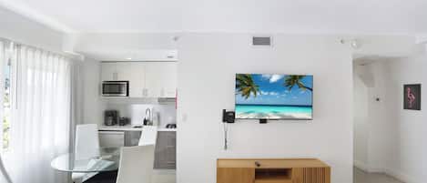 A modern living room with a glass dining table, white chairs, a wooden console table, and a mounted TV displaying a beach scene.
