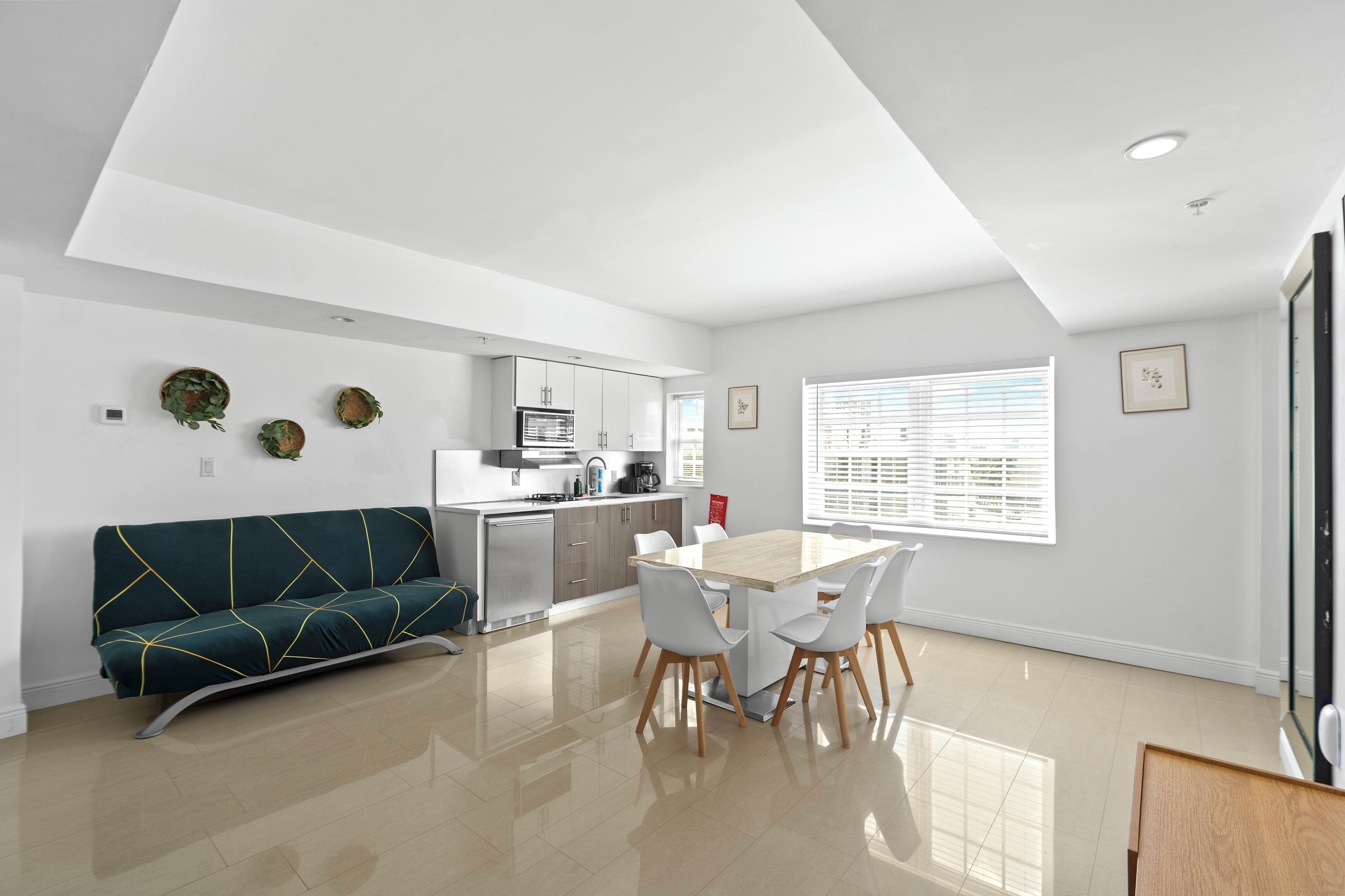 A modern kitchen with white cabinets, a dining table with chairs, a green sofa, and a large window with blinds.