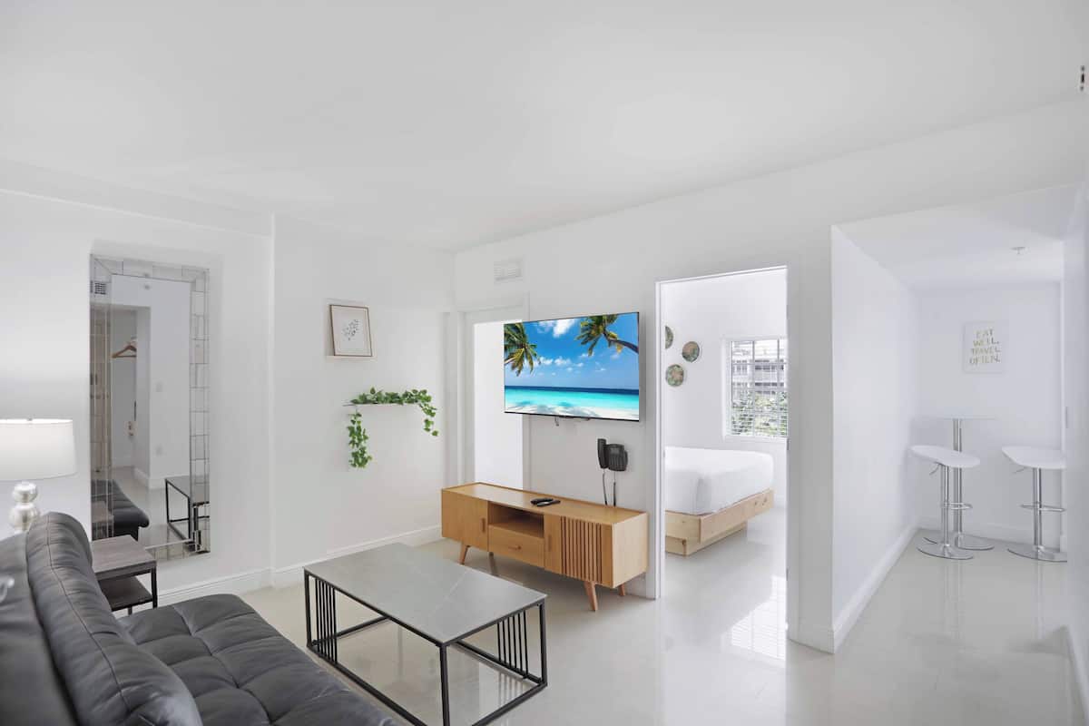 A modern living room with a sofa, coffee table, TV, and a bed in the adjacent room.