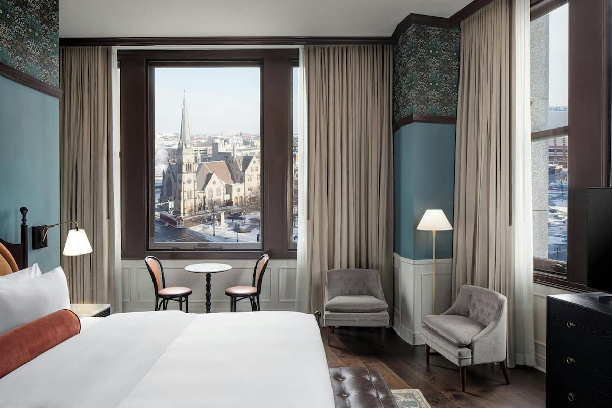 A hotel room with a large bed, two armchairs, a small table, and a view of a cityscape through a large window.