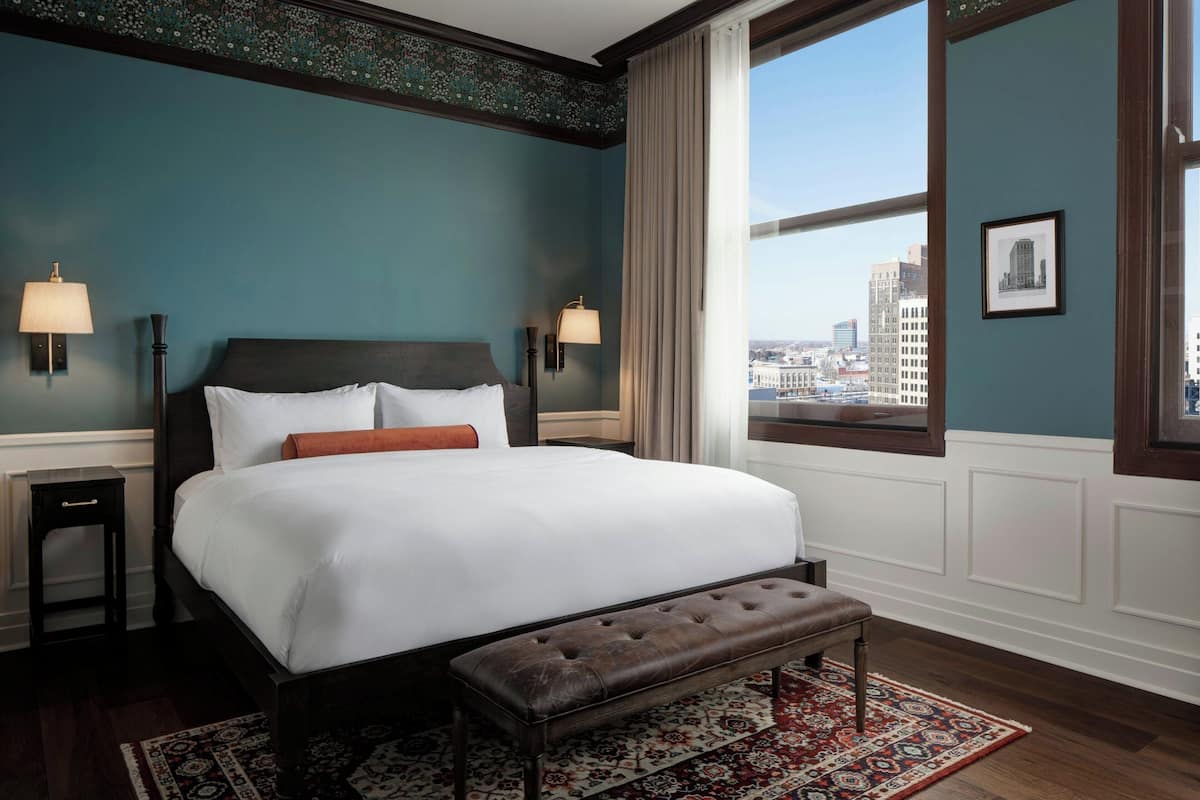 A hotel room with a large bed, two bedside lamps, a bench, a patterned rug, and a city view through a large window.