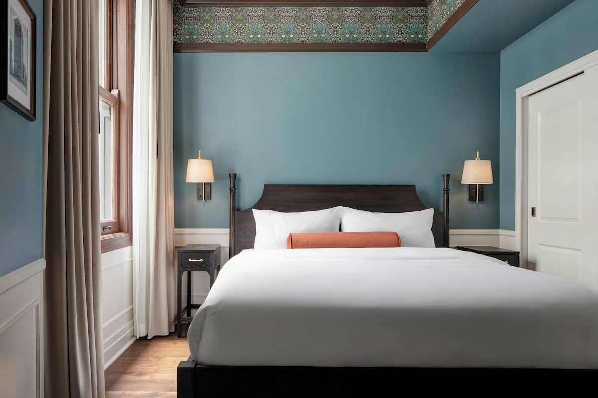 A neatly made bed with white linens and a single orange accent pillow, flanked by two bedside tables with lamps, in a room with blue walls and a patterned ceiling.