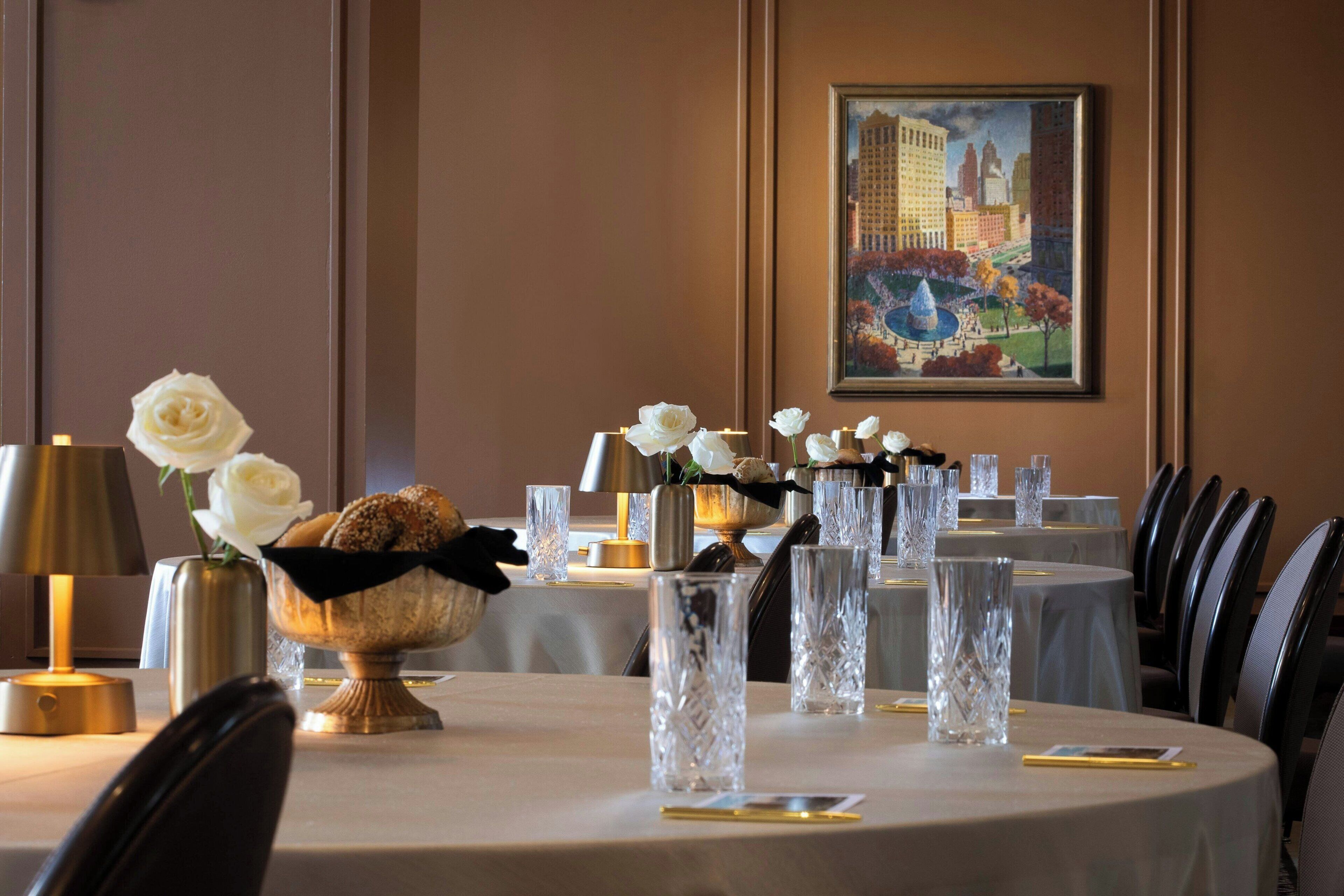 A dining area with round tables, white roses, crystal vases, and a framed painting on the wall.