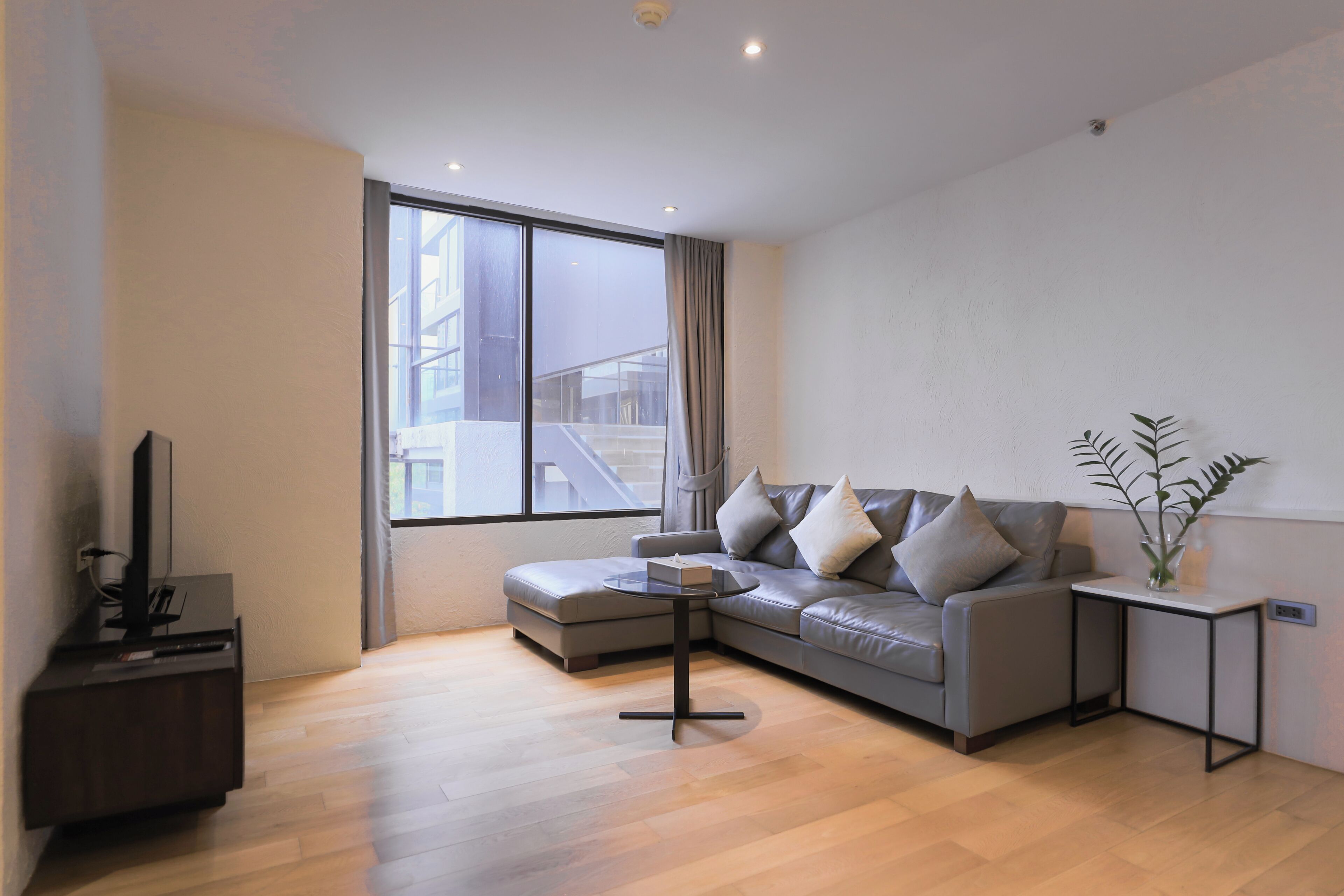 Een moderne woonkamer met een grijze bank, een klein bijzettafeltje, een flatscreen televisie en een groot raam.